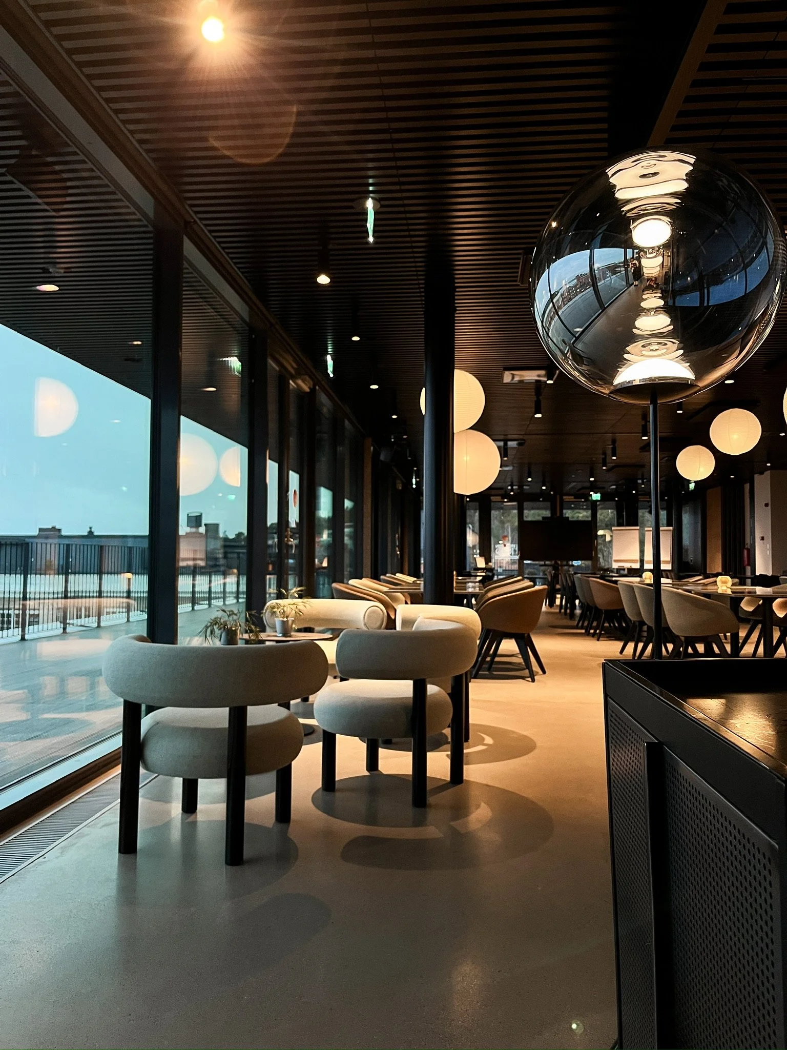 Interior of a modern restaurant or cafe with round hanging pendant lights, large glass windows, beige and white chairs, and a black ceiling with wooden slats.