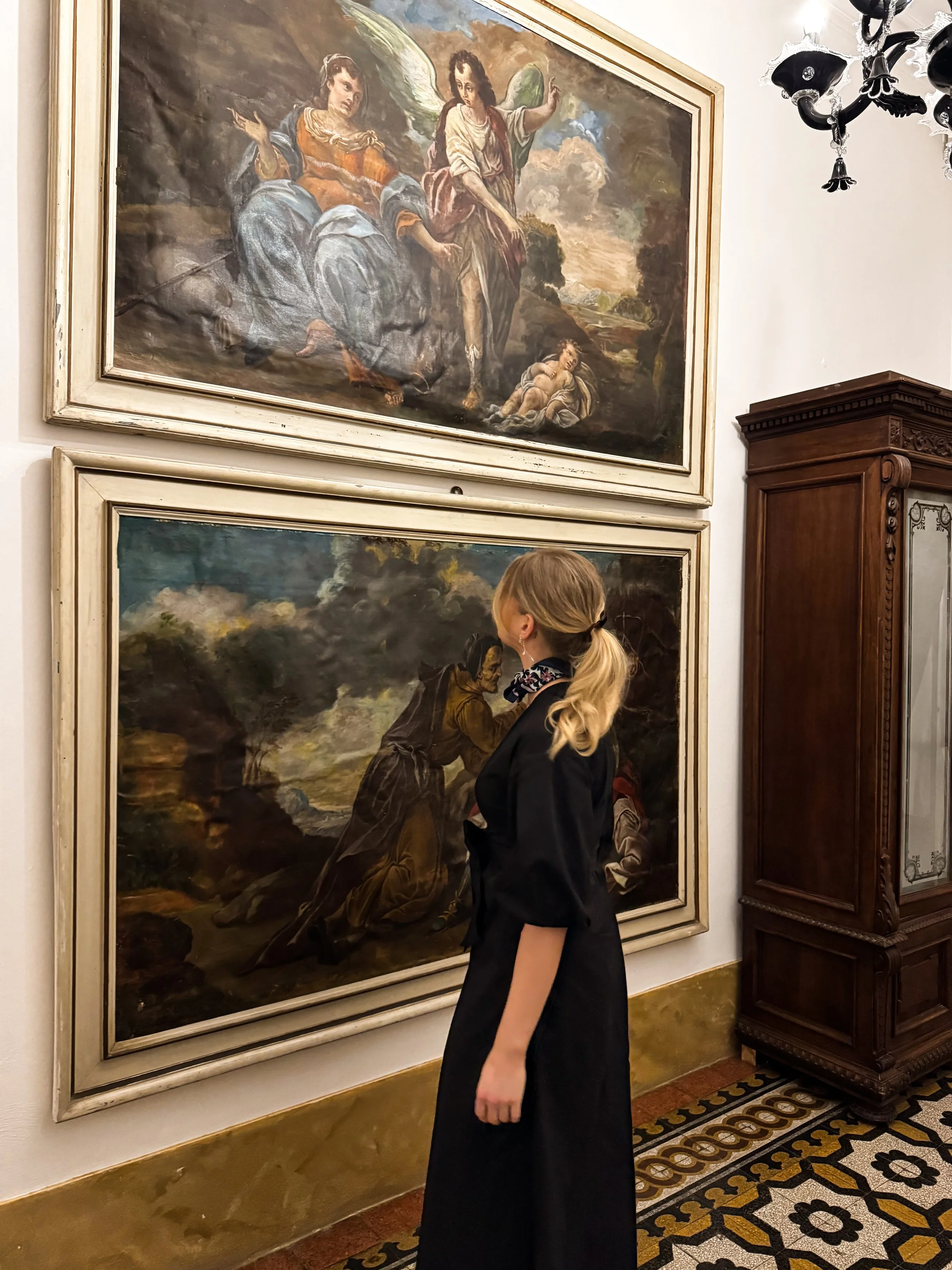 A woman with blonde hair tied back, wearing a black dress and a patterned scarf, standing in front of two large framed classical paintings in an art gallery.