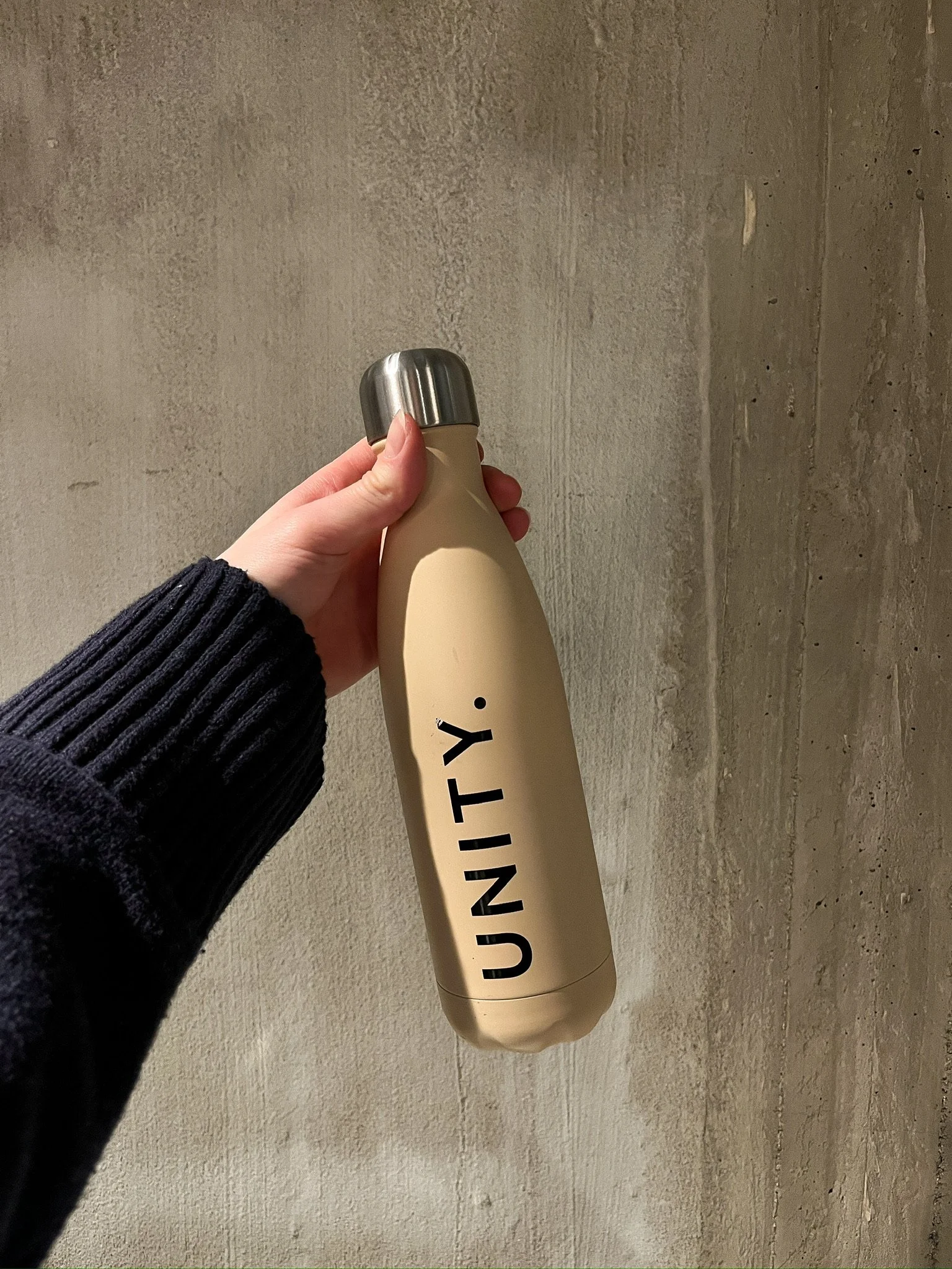 A person holding a beige insulated water bottle with a silver cap, labeled 'UNITY' in black letters, against a textured concrete wall background.