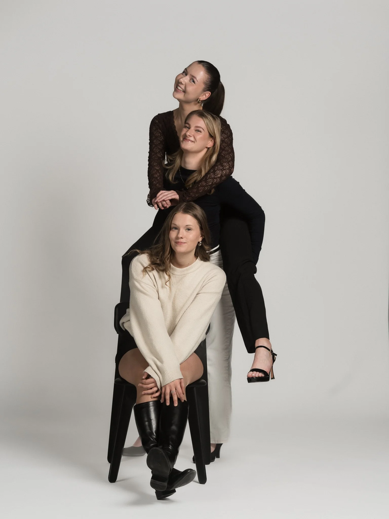 Three women posing in a studio with a plain background. The woman in the front is sitting on a chair, wearing a cream sweater and black boots. The woman in the middle is sitting on the lap of the woman at the back, wearing a black top and pants. The woman at the back is standing, wearing a black dress with lace sleeves and high heels.