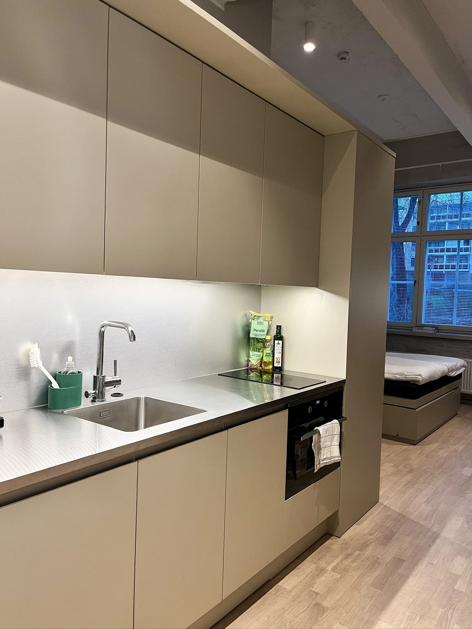 Modern kitchen with beige cabinets, stainless steel sink, bottle of soap, green toothbrush holder, and stovetop. In the background, a bedroom with a bed and large window.