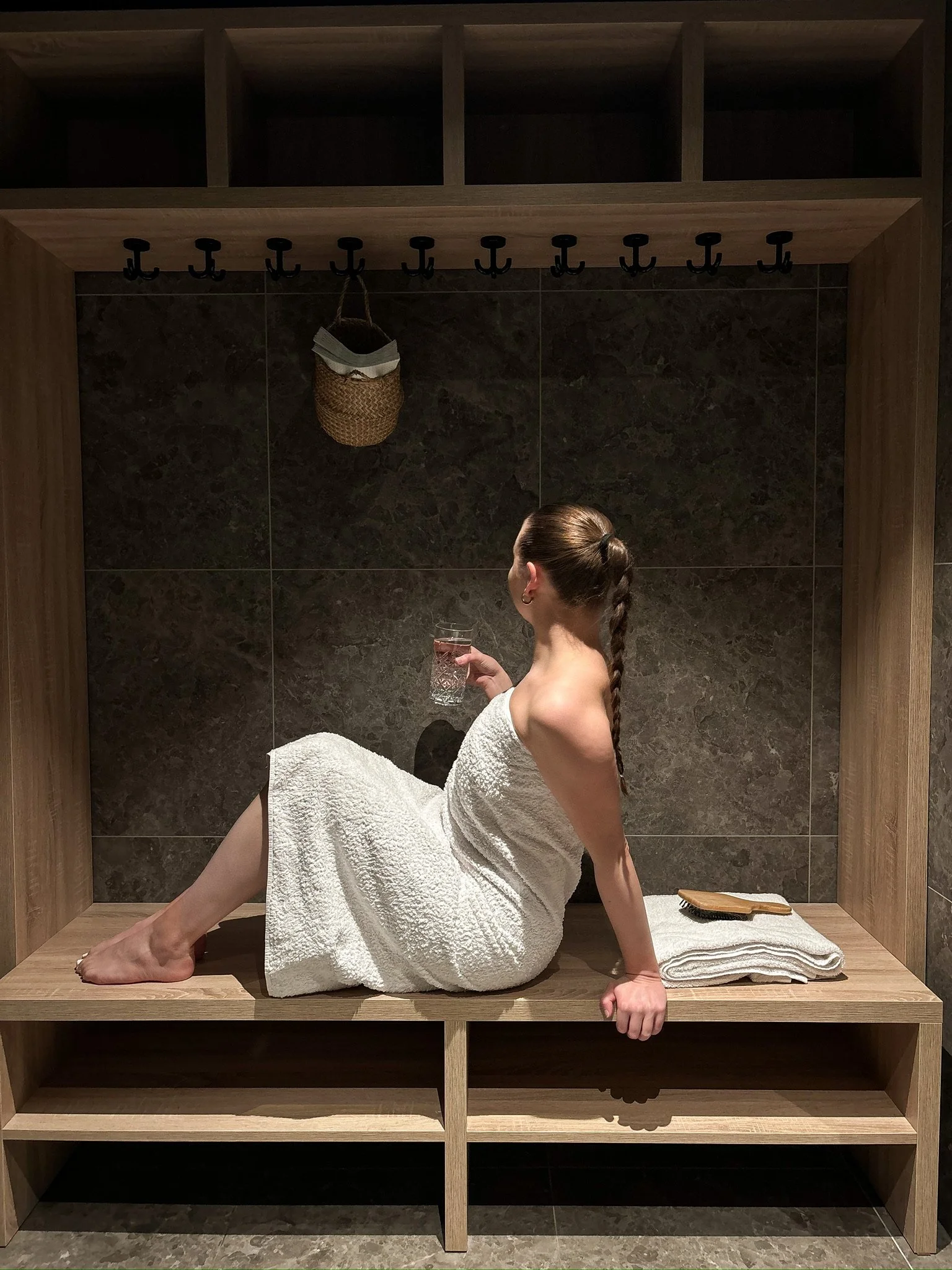 A woman with a braid, wrapped in a white towel, sitting on a wooden bench in a steam room or sauna, holding a glass of water, with additional towels and grooming brush nearby.