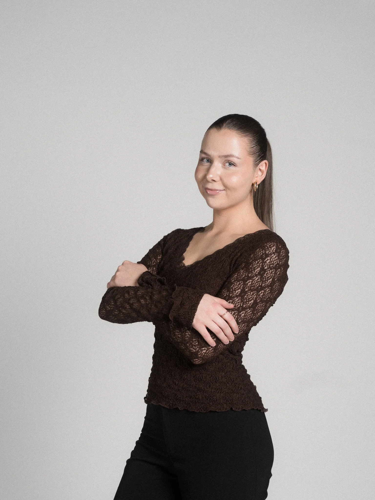 A young woman with long brown hair in a high ponytail, wearing a black, long-sleeved lace top and black pants, stands with arms crossed, smiling softly against a plain light gray background.
