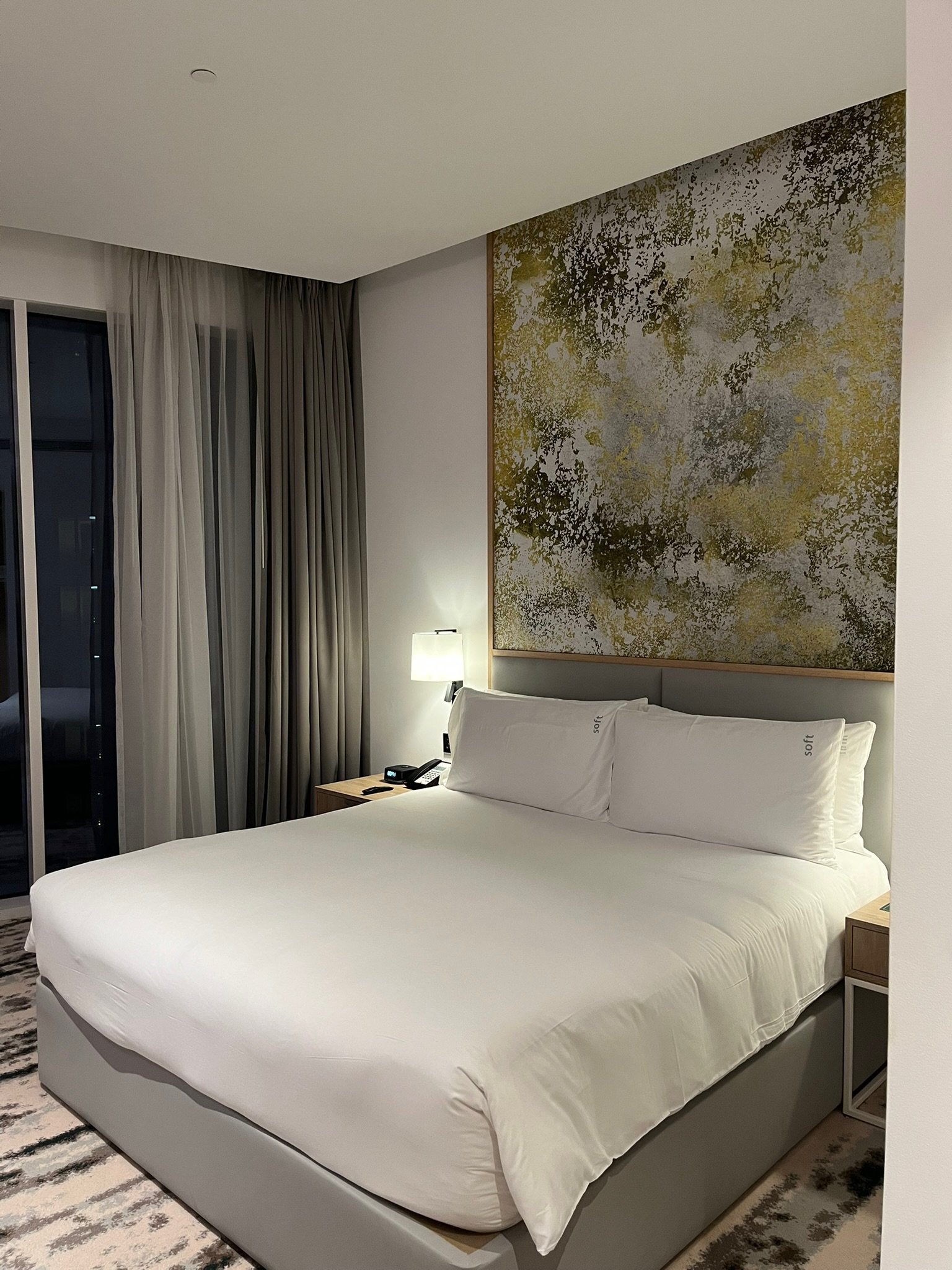A neatly made hotel bed with white bedding, pillows, and a headboard, in a modern hotel room with wall art, bedside lamps, a phone, and curtains beside a sliding glass door