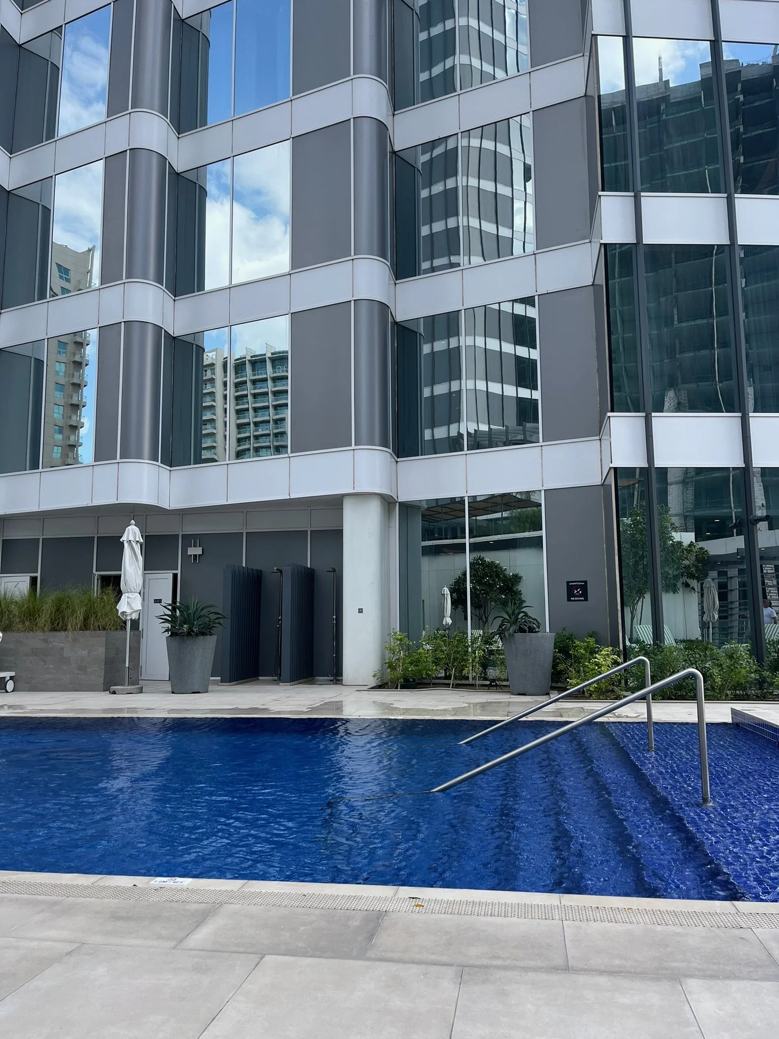 A modern building with reflective glass windows overlooking a swimming pool with a blue tiled bottom. There are potted plants near the building and a couple of closed umbrellas on the patio.