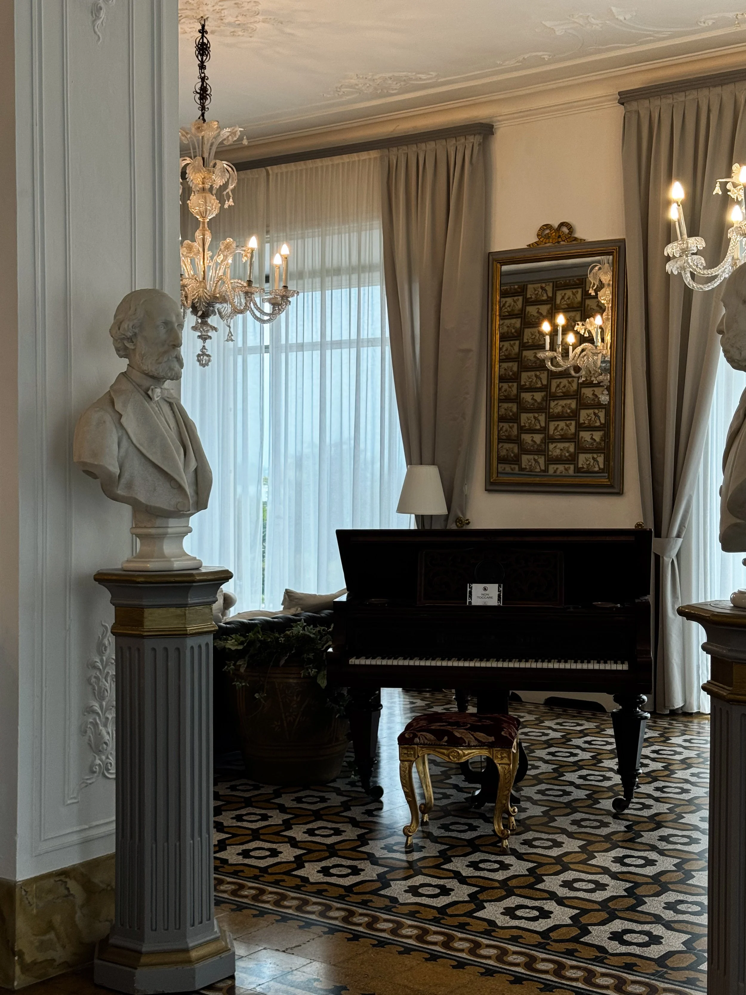 Elegant room with chandeliers, a grand piano, marble busts on pedestals, and patterned tile flooring.
