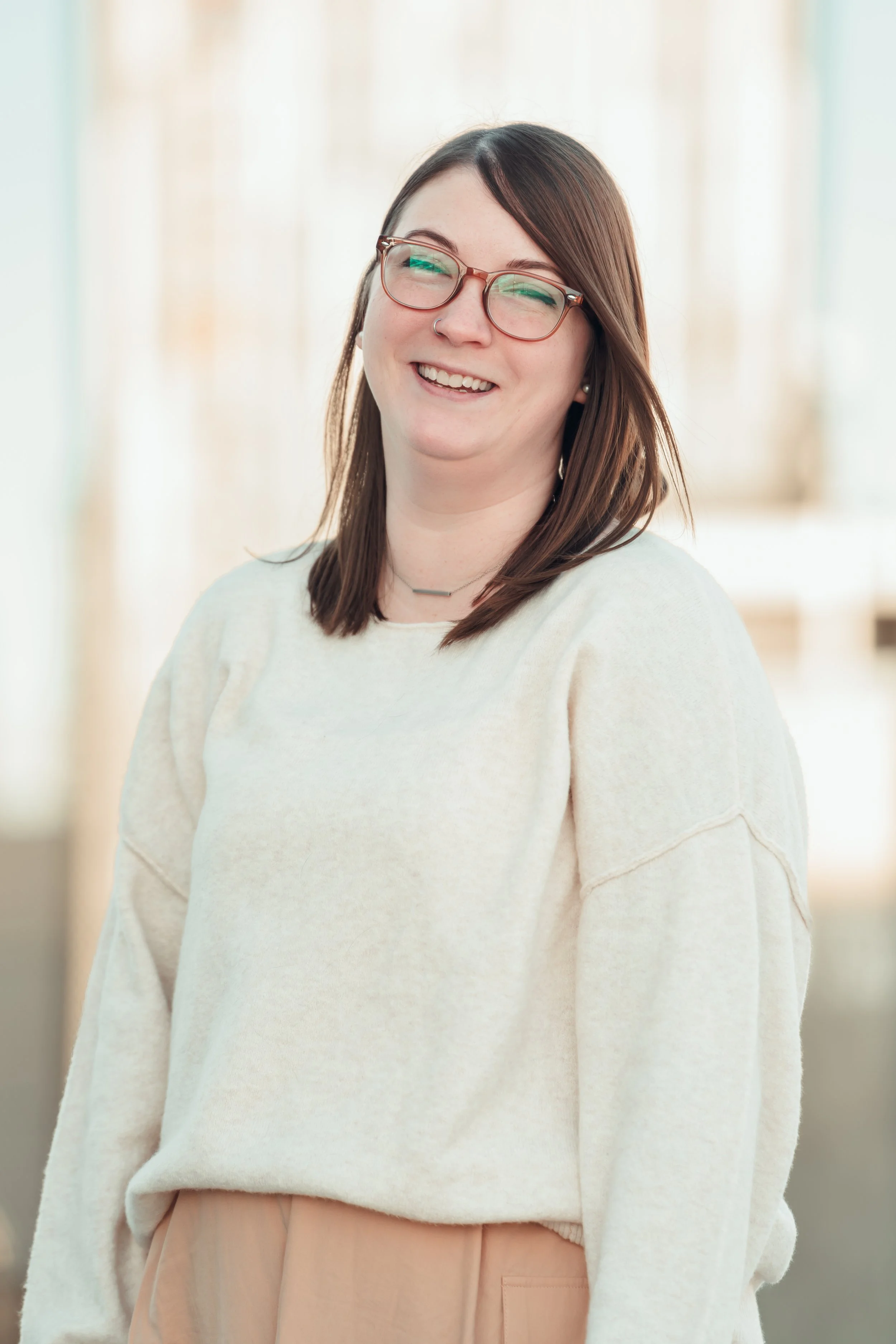 A smiling woman with shoulder-length brown hair, wearing glasses, a white sweater, and beige pants, standing indoors with blurred background.
