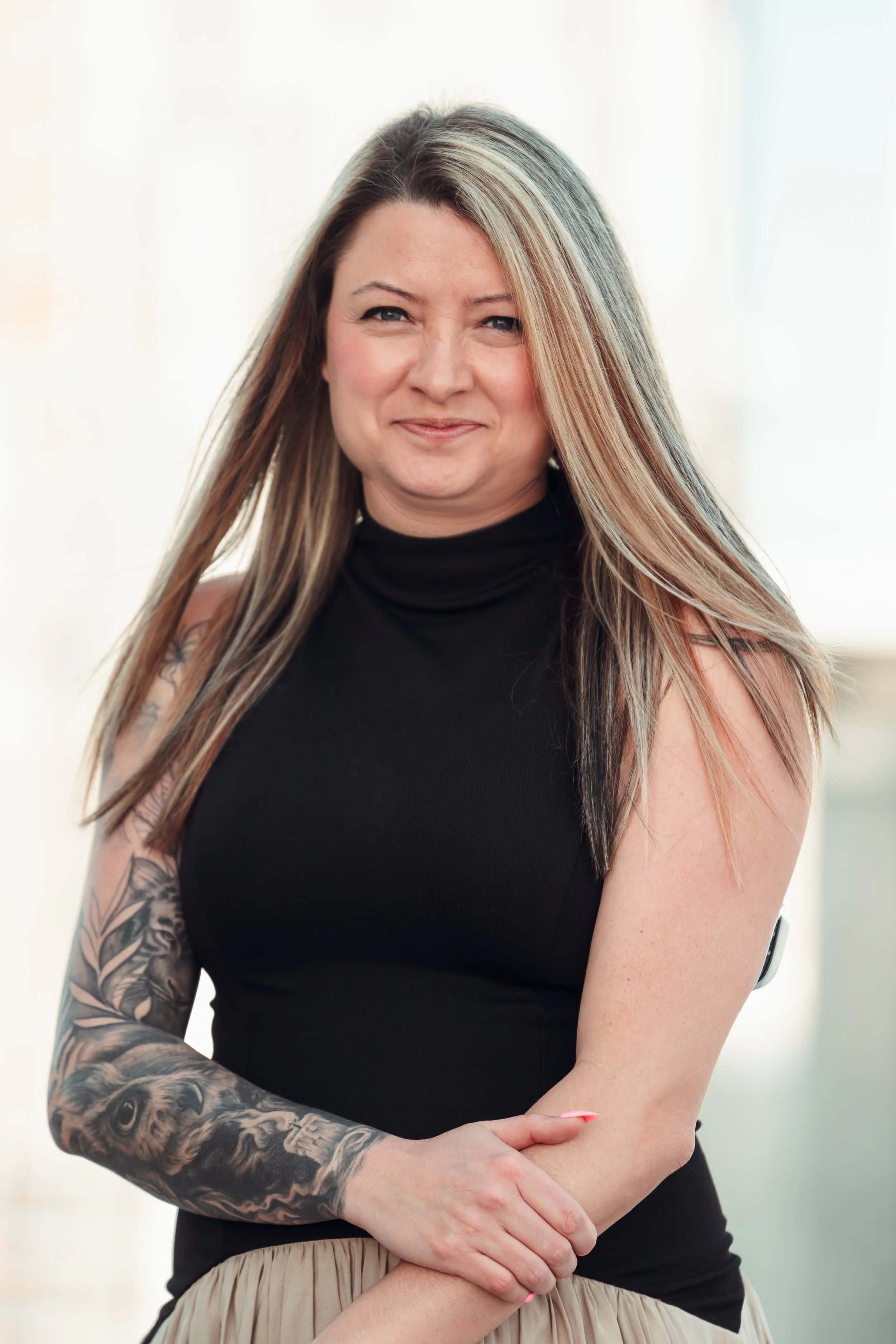 A woman with long blonde hair, wearing a black sleeveless top and a beige skirt, standing indoors with a neutral background.