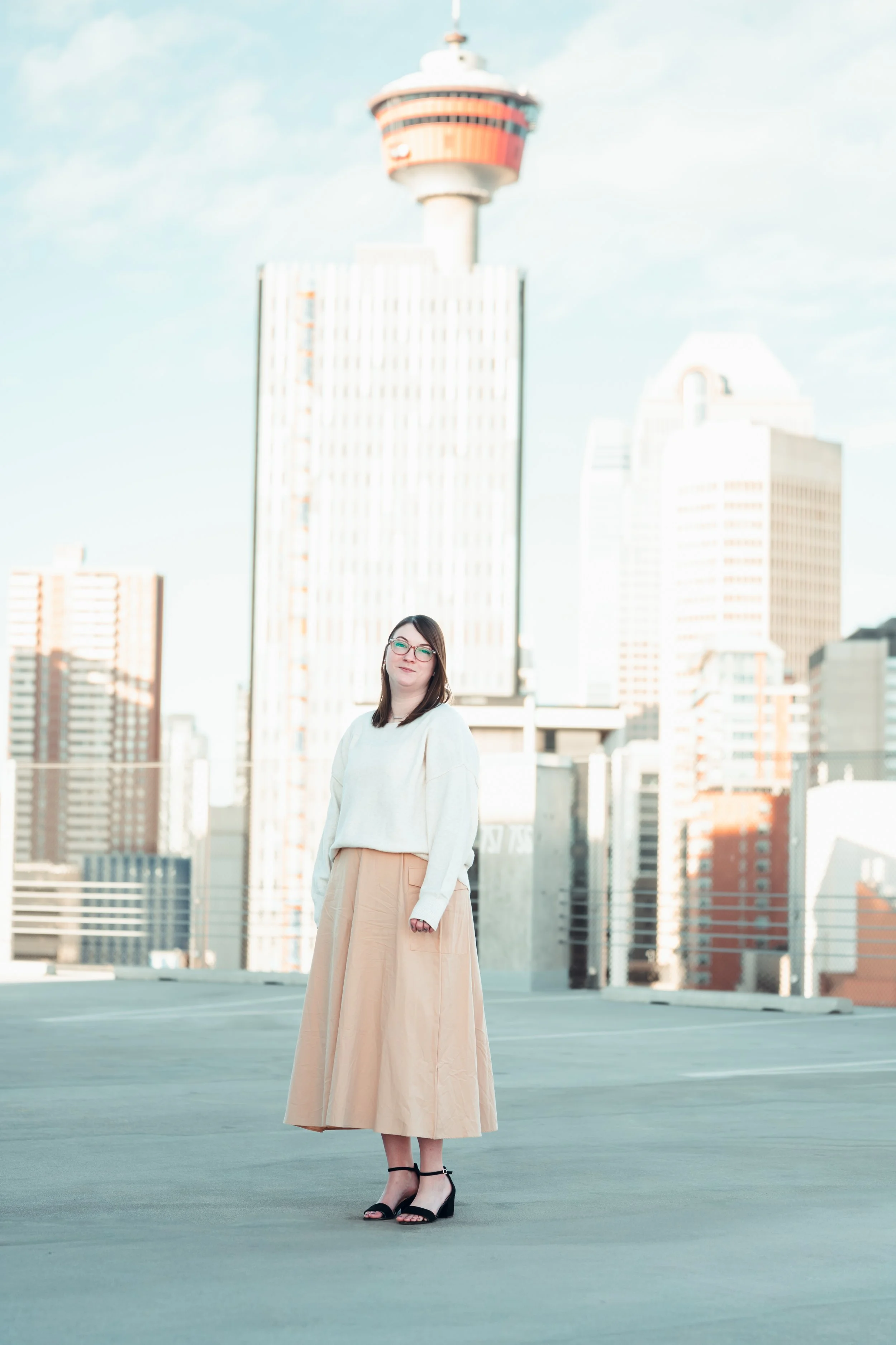 A woman standing on a rooftop parking lot with a city skyline and the Calgary Tower in the background, wearing a beige skirt, white sweater, and black sandals.