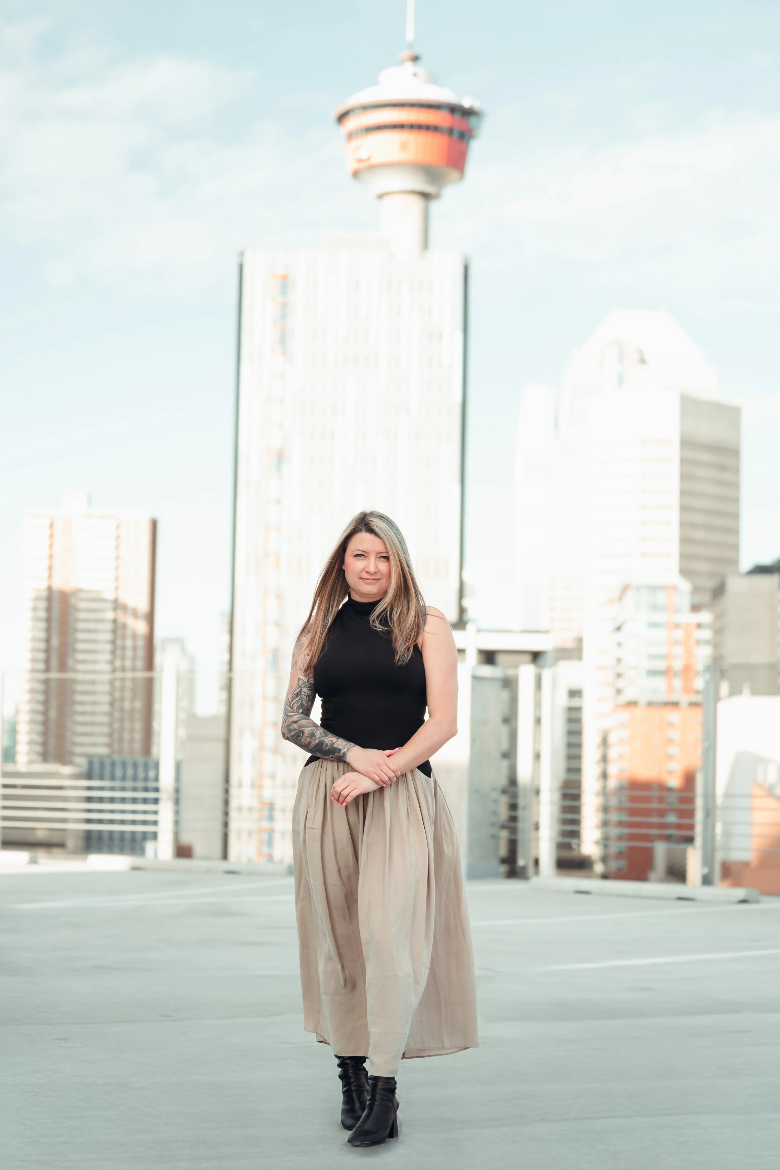 A woman with long hair wearing a black sleeveless top, beige wide-leg pants, and black boots stands on a rooftop with a city skyline and a communications tower in the background.