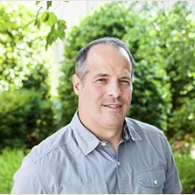 A man with short dark hair and a receding hairline, wearing a light blue collared shirt, standing outdoors in front of greenery and smiling. Jim Kupec