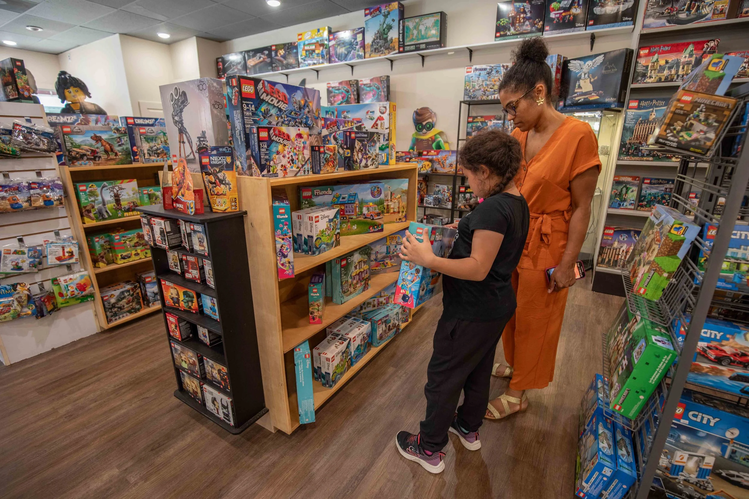 A woman and a young girl shopping for LEGO sets in a toy store, standing near shelves filled with various LEGO and other toy sets.