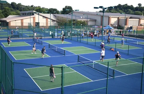 Multiple groups of people playing tennis on blue and green courts at a sports complex with a chain-link fence around the perimeter.