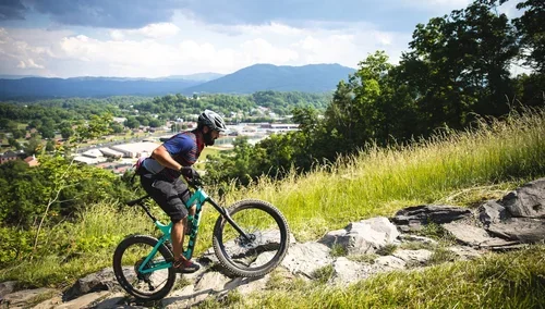 A person riding a mountain bike uphill on a grassy trail with rocks, trees, and a cityscape with mountains in the background.