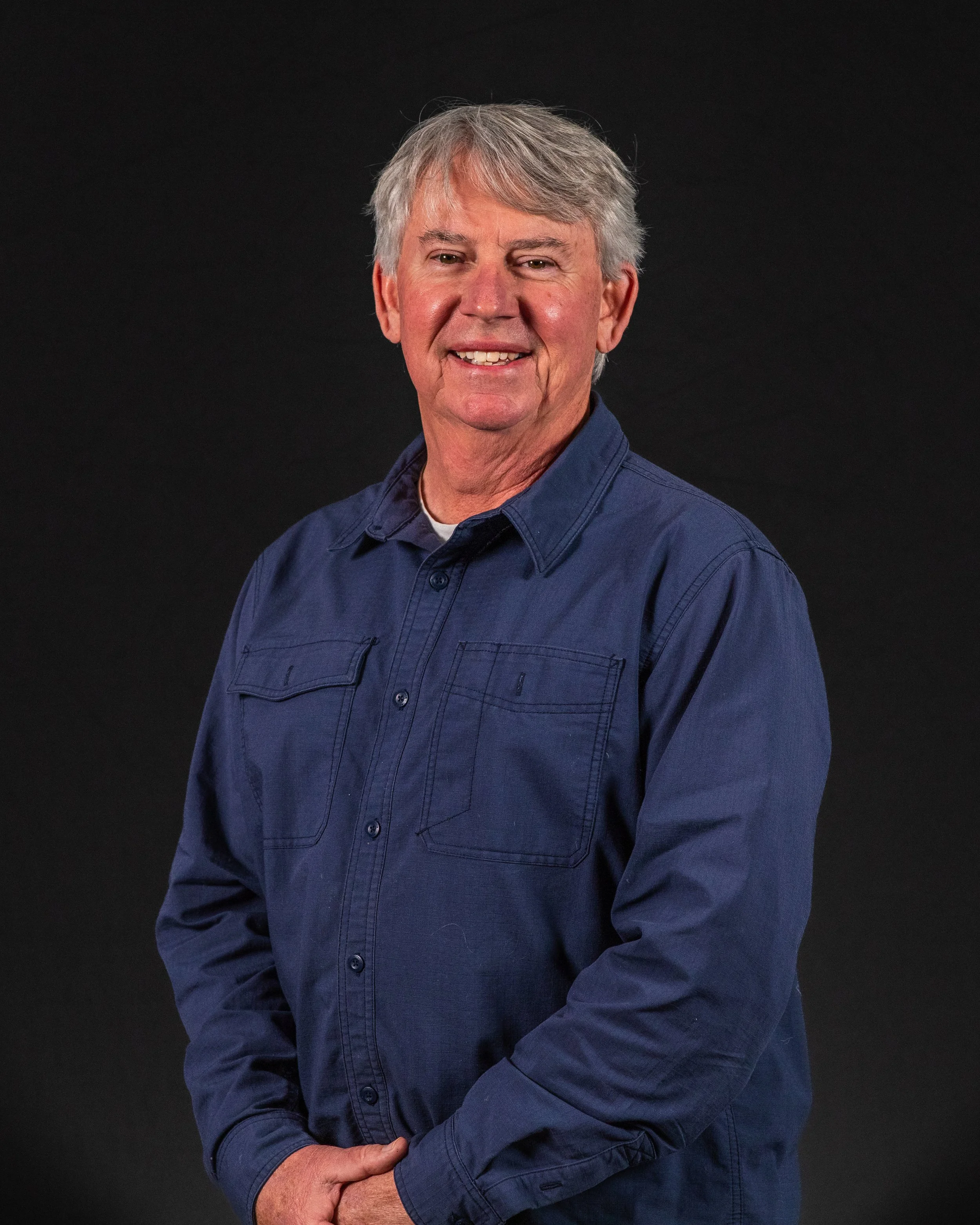 A smiling middle-aged man with gray hair, wearing a blue button-up shirt, standing against a black background.