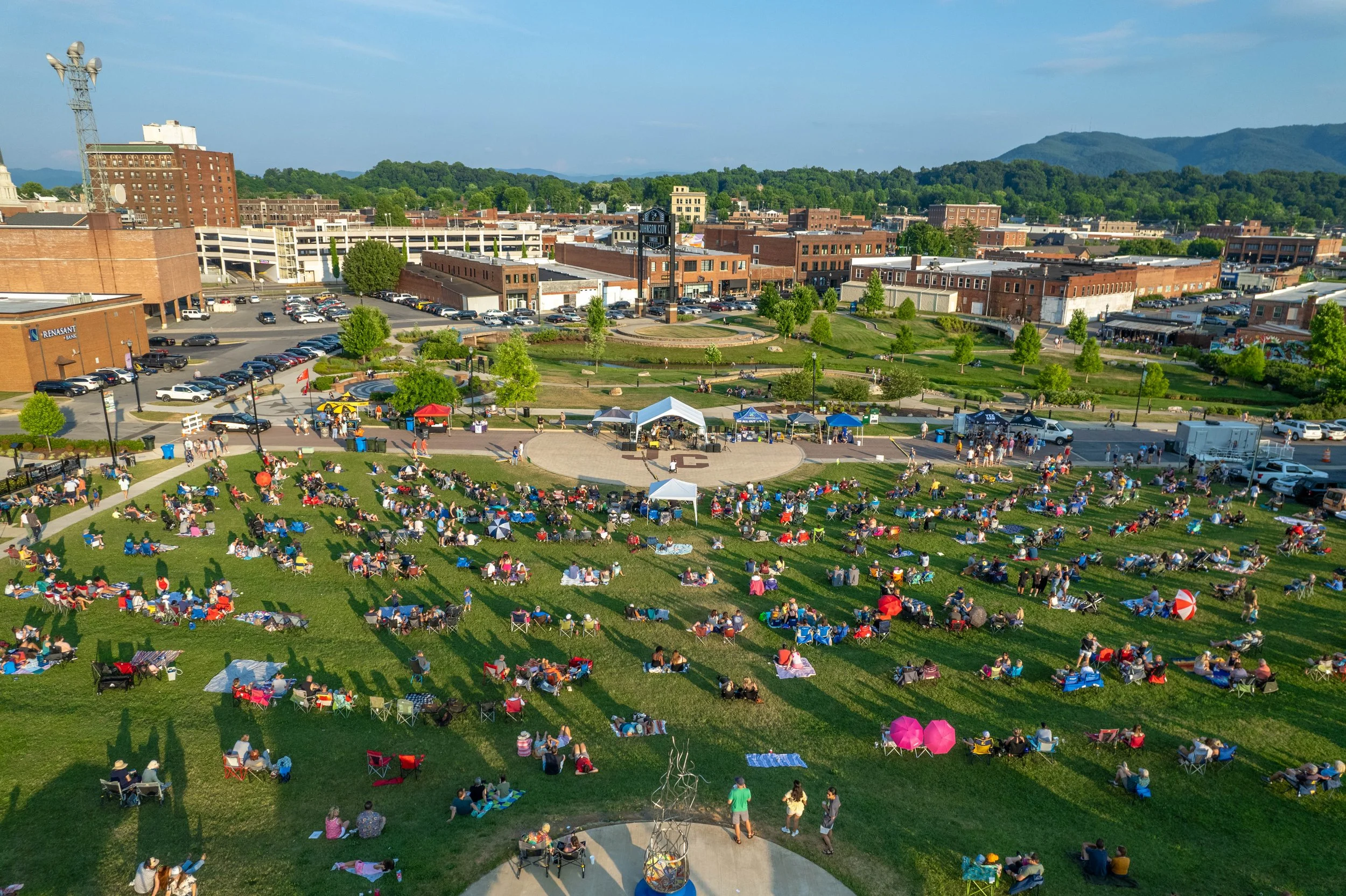 An outdoor concert in a park with many people sitting on blankets and chairs, tents set up, and a stage, with a cityscape and hills in the background.
