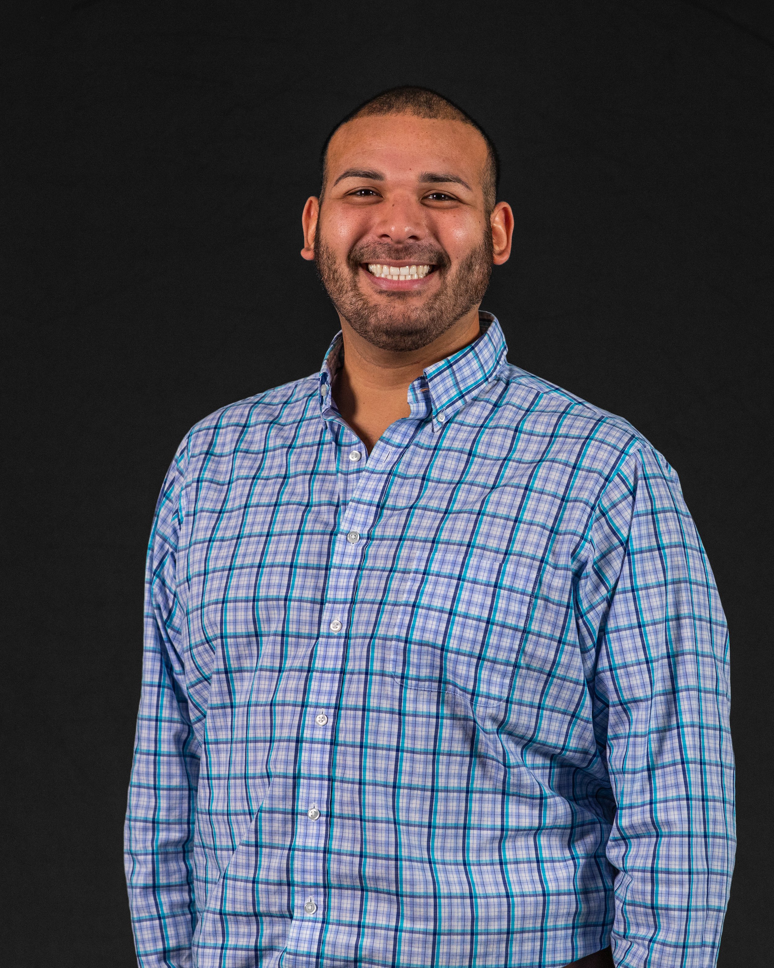 A man with a beard wearing a blue plaid button-down shirt, smiling at the camera, standing against a black background.