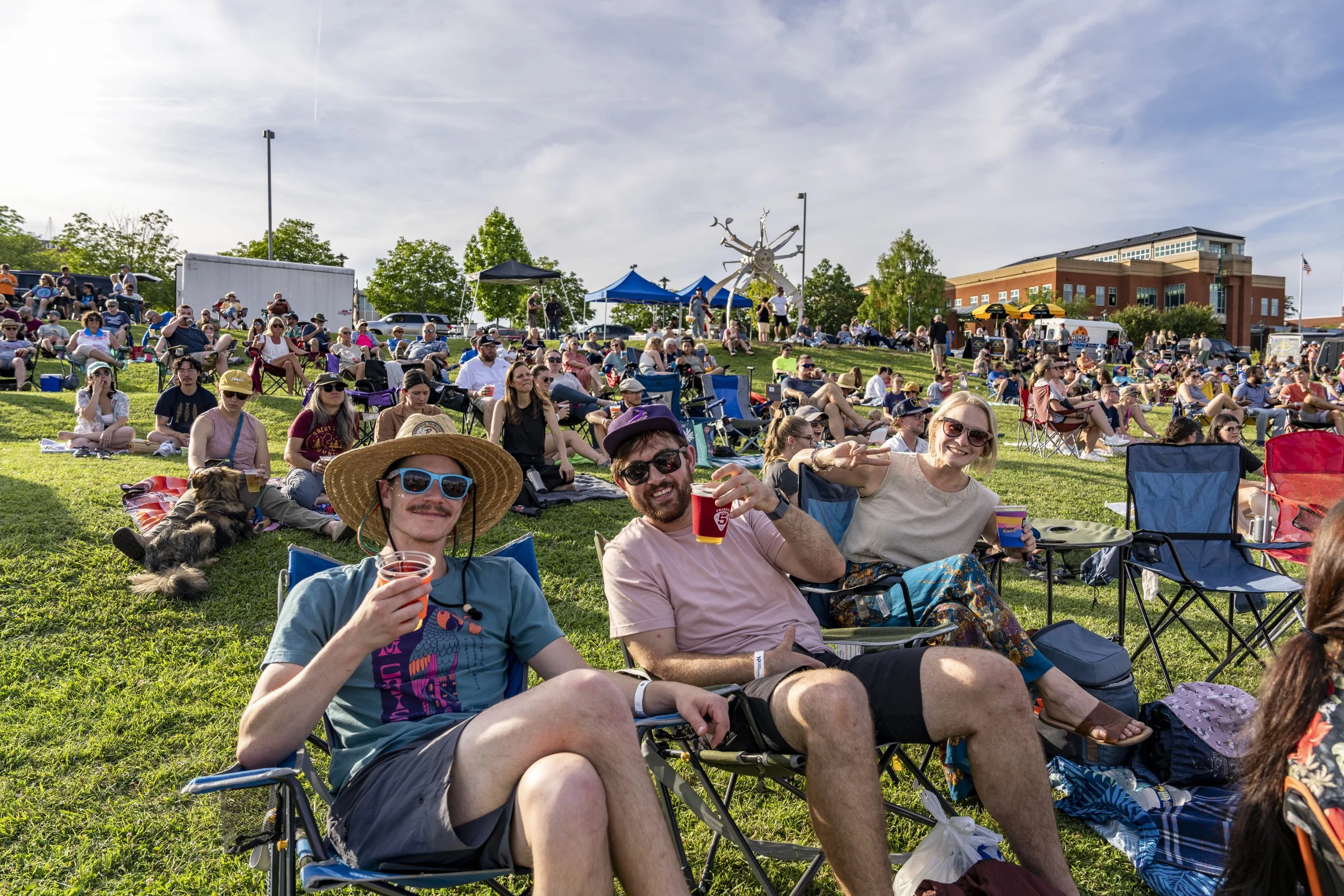 People attending an outdoor event on a grassy hill, sitting on chairs and blankets, with some holding drinks, under a partly cloudy sky.