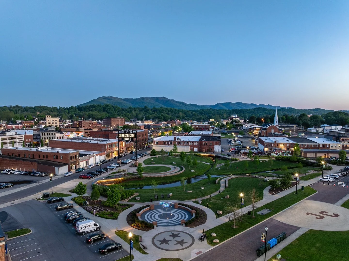 Aerial view of Johnson City, Tennessee at dusk, showing a park with fountain, green spaces, a parking lot, historic brick buildings, a church, and distant mountains.