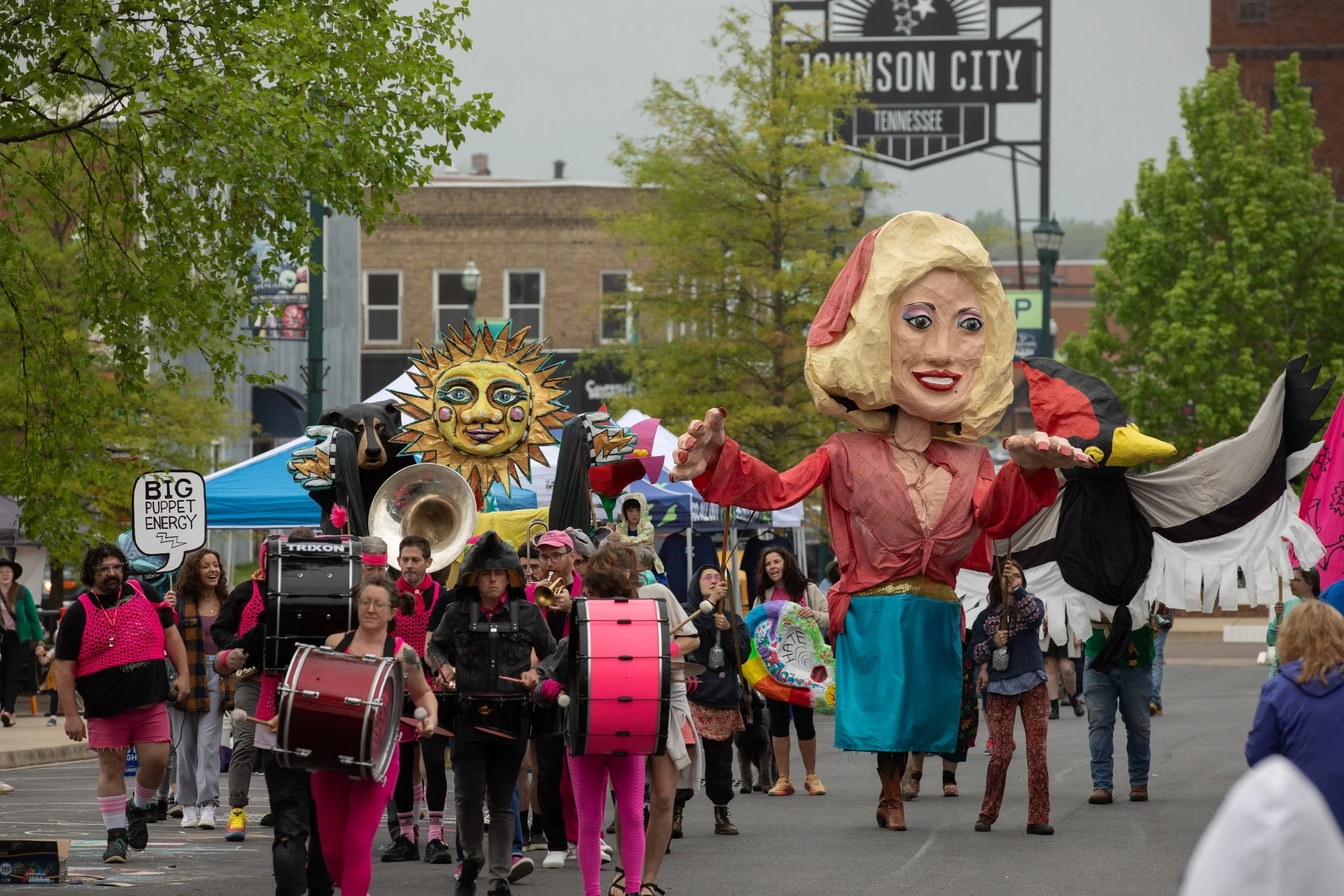 A parade with people holding large puppets, including a woman with a big head and a bird, with a sun puppet, musicians playing drums and instruments, and colorful costumes in an urban area with trees and buildings in the background.