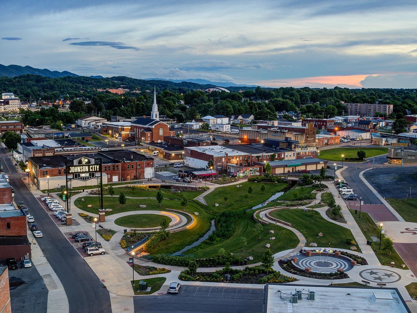 A view of downtown Johnson City, Tennessee, at dusk, featuring a park with winding paths, a small stream, and a circular seating area with the Tennessee state emblem. Surrounding buildings include a church, shops, and parking areas with cars, with mountains in the background and a partly cloudy sky.