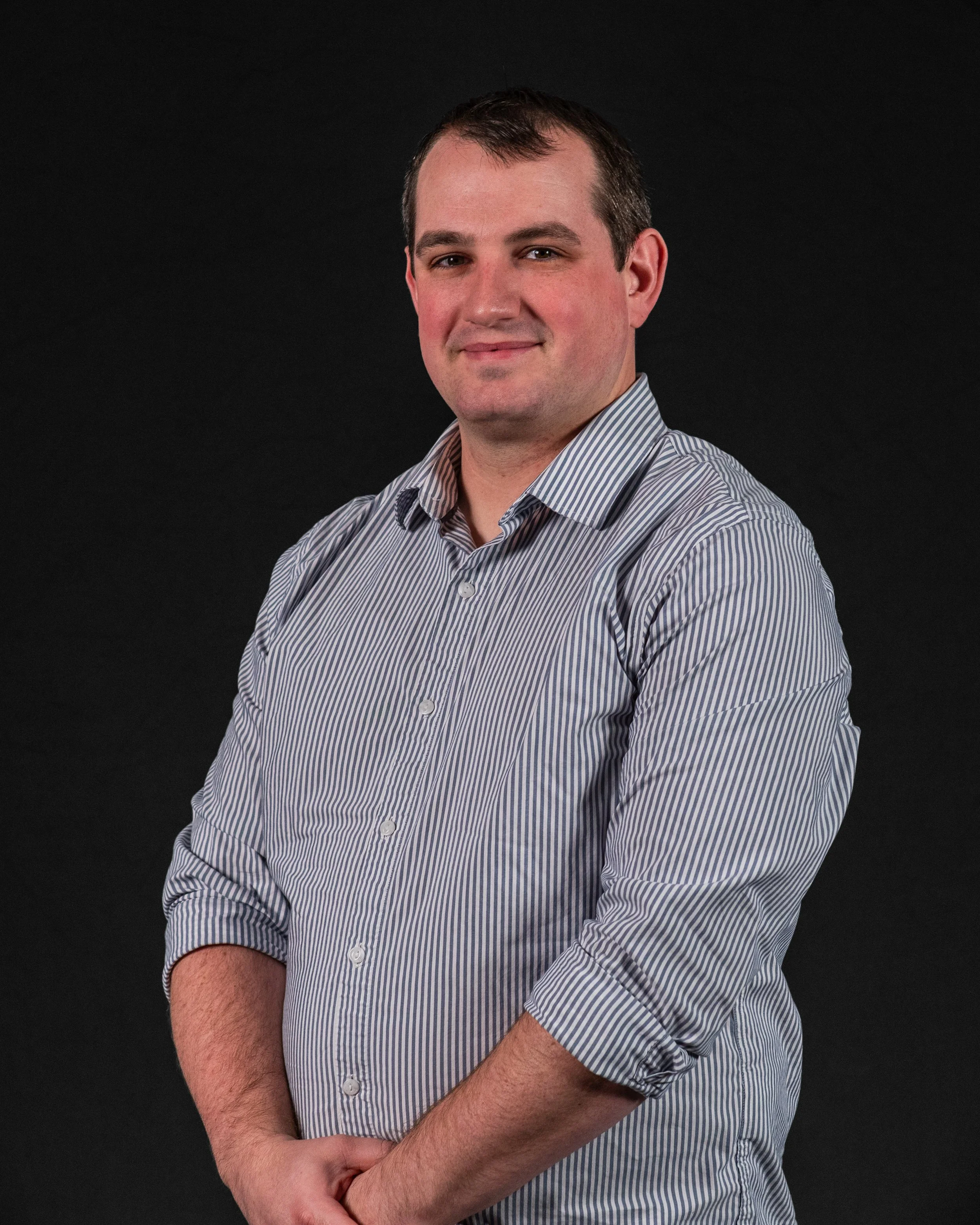 A young man with short dark hair and fair skin wearing a light-colored, striped button-up shirt poses against a black background.