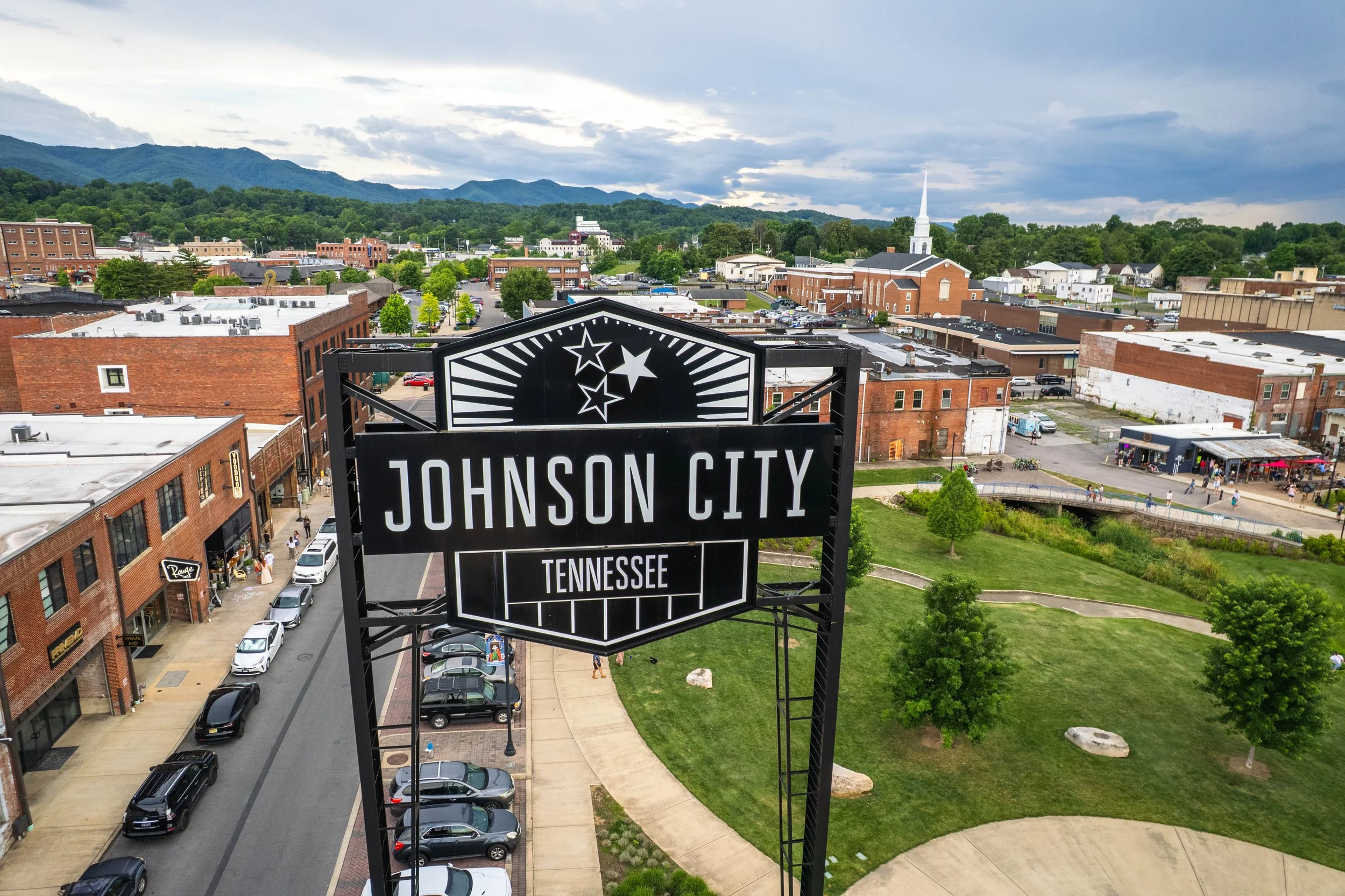 View of Johnson City, Tennessee, downtown area with a large sign in the foreground, brick buildings, parking lots, people walking, green park with trees, and distant mountains under a cloudy sky.