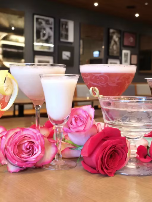 Four cocktails with pink, white, and clear liquids and a pink flower, surrounded by pink and red roses on a wooden table in a restaurant or bar setting.