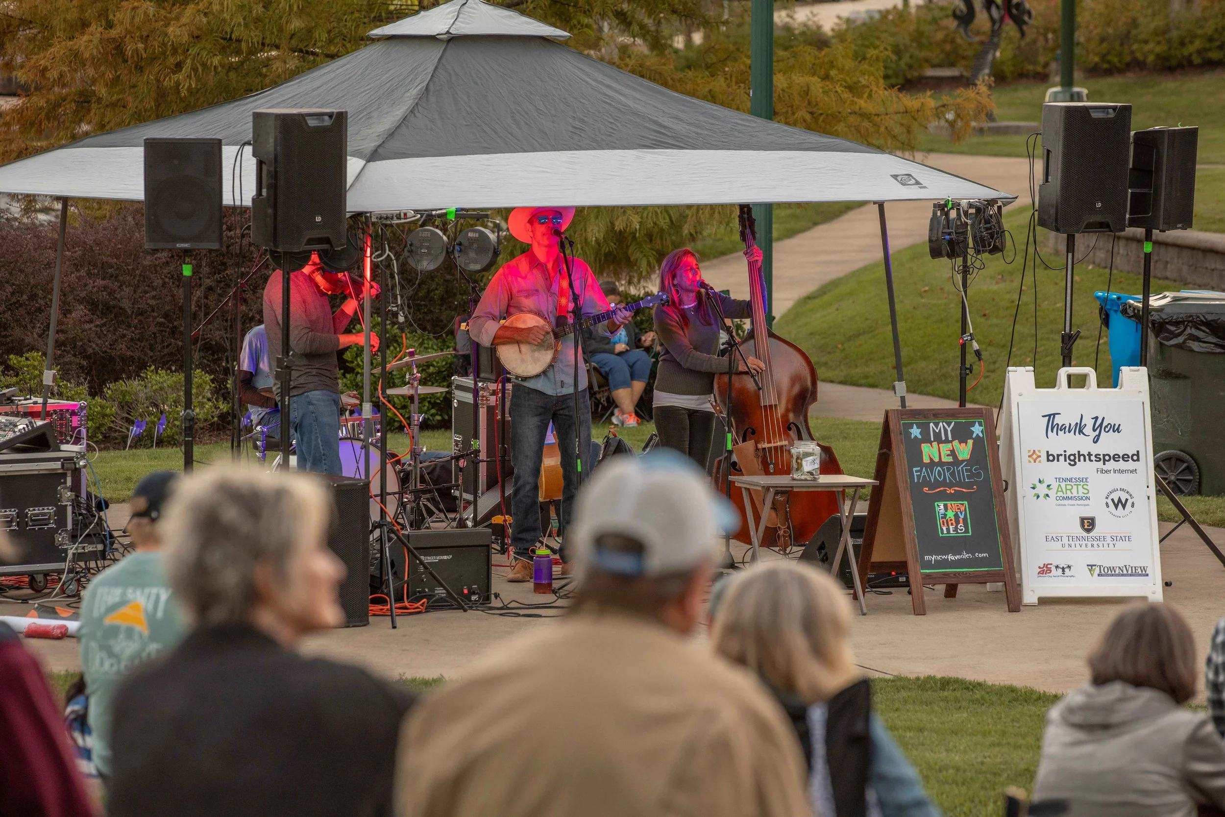 Community outdoor concert with a band playing under a canopy, audience seated on grass, and signs for sponsors.