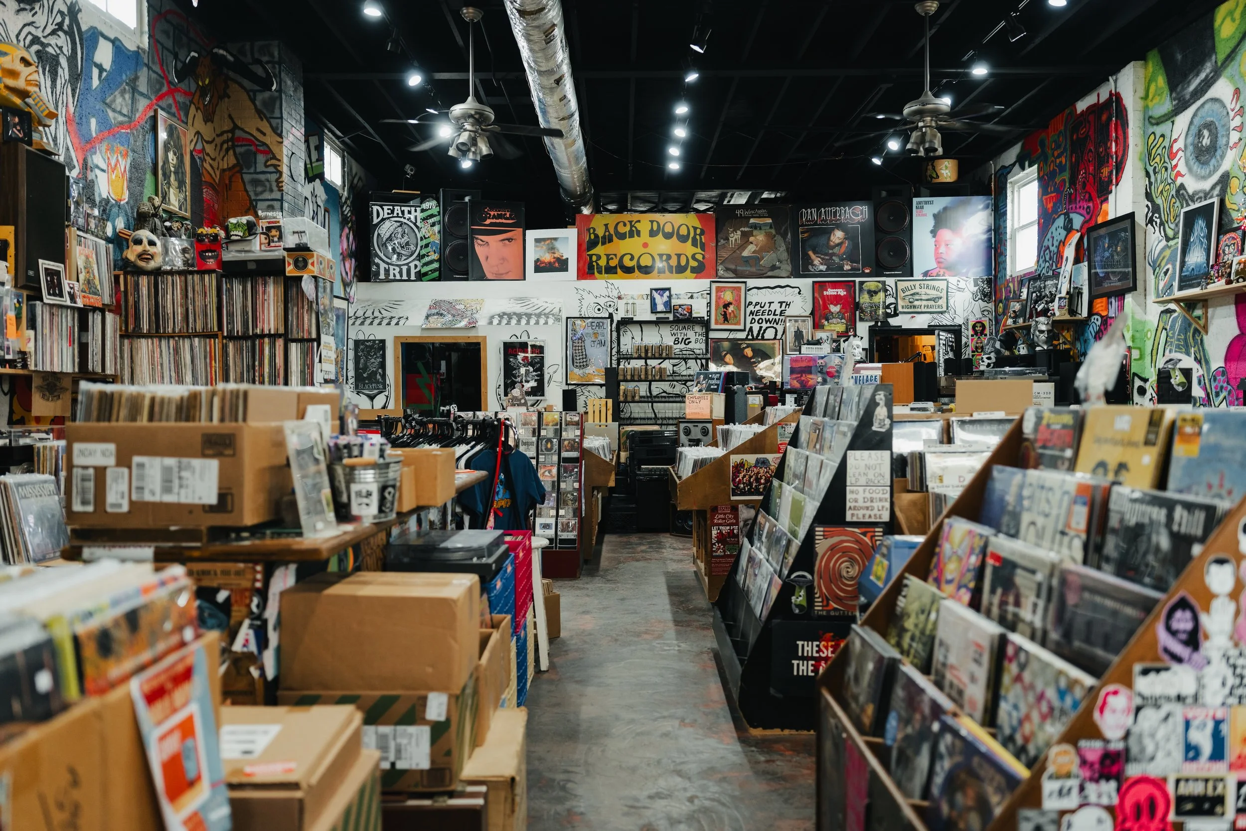 Interior of a record store filled with vinyl records, posters, and artwork on the walls, with aisles of boxes and shelves of records.