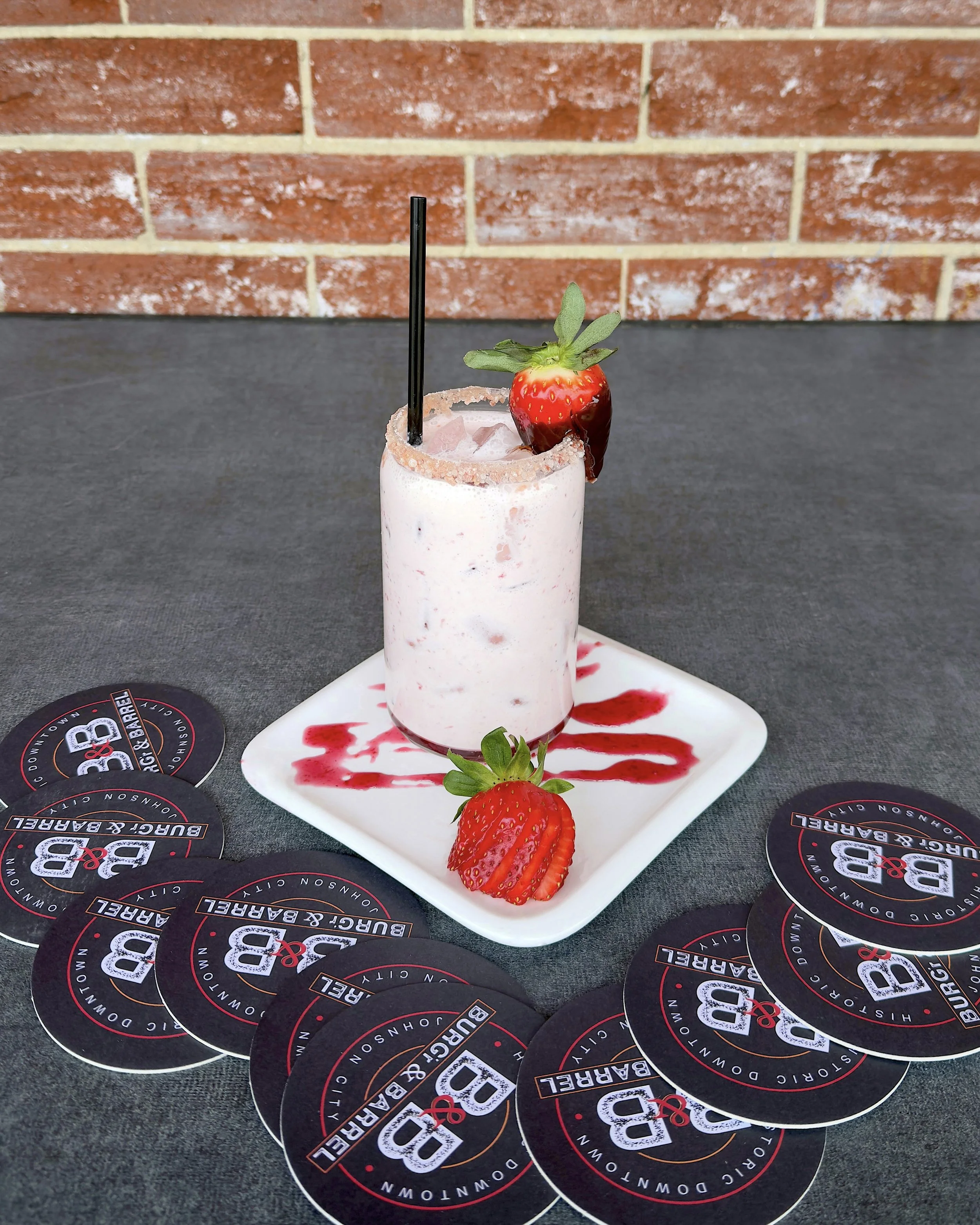 A strawberry milkshake in a glass with a salted rim, garnished with a strawberry, served on a white plate with strawberry syrup. Multiple coasters with the logo 'B & B' surround the plate on a dark surface, against a brick wall background.