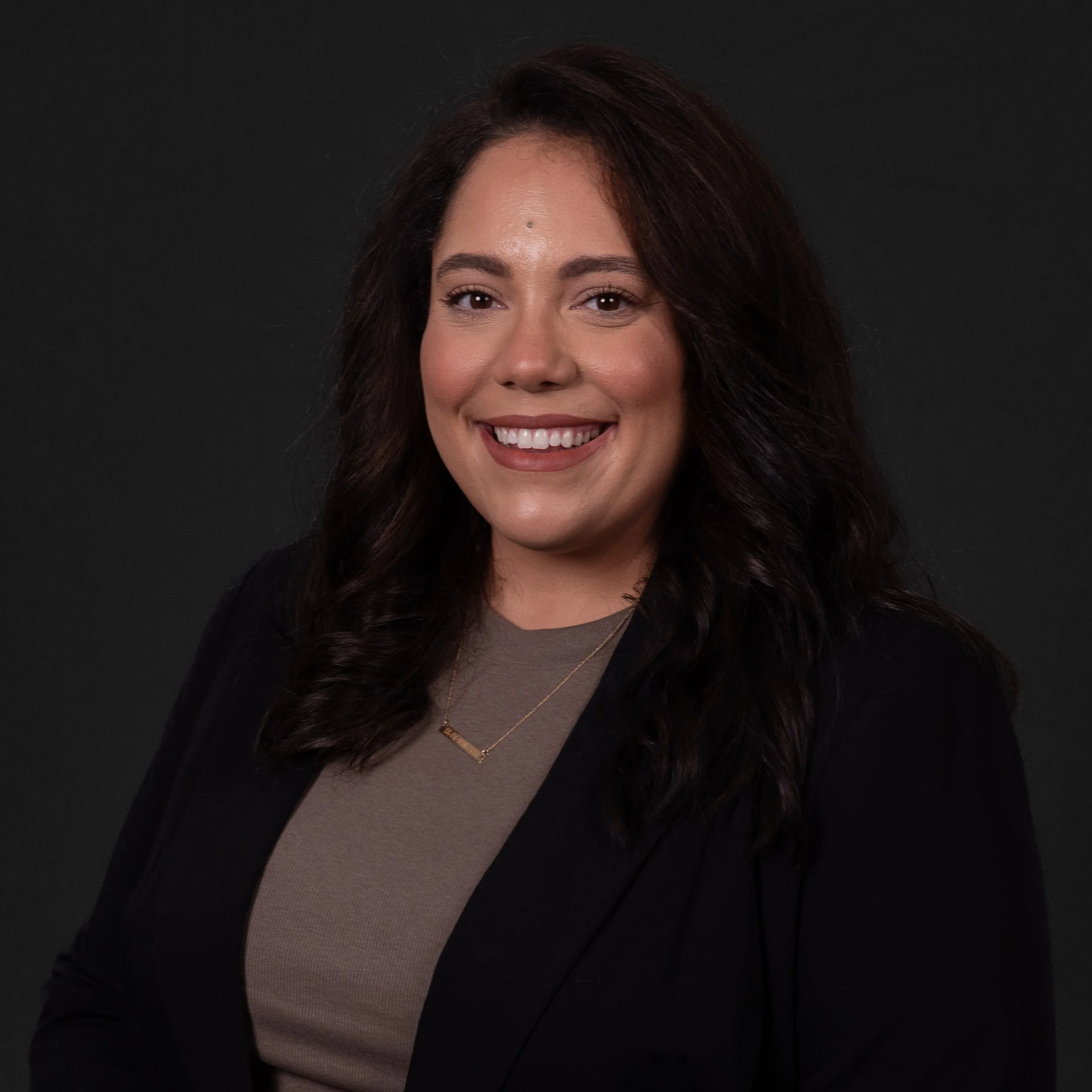A woman with dark wavy hair smiling, wearing a beige top and black blazer, against a dark background.