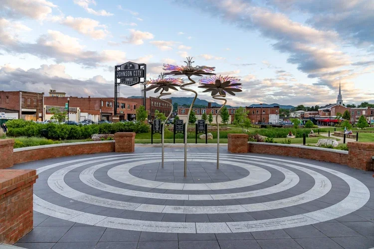 Public plaza in Johnson City with a spiral pattern on the ground, floral sculpture, and brick wall borders, with buildings, greenery, and a church spire in the background.