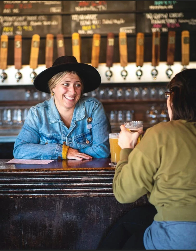 Two women talking at a bar with beers. One woman is smiling, wearing a denim jacket and a wide-brimmed hat. The other woman has glasses and is wearing a green jacket. The background shows beer taps and a menu on the wall.