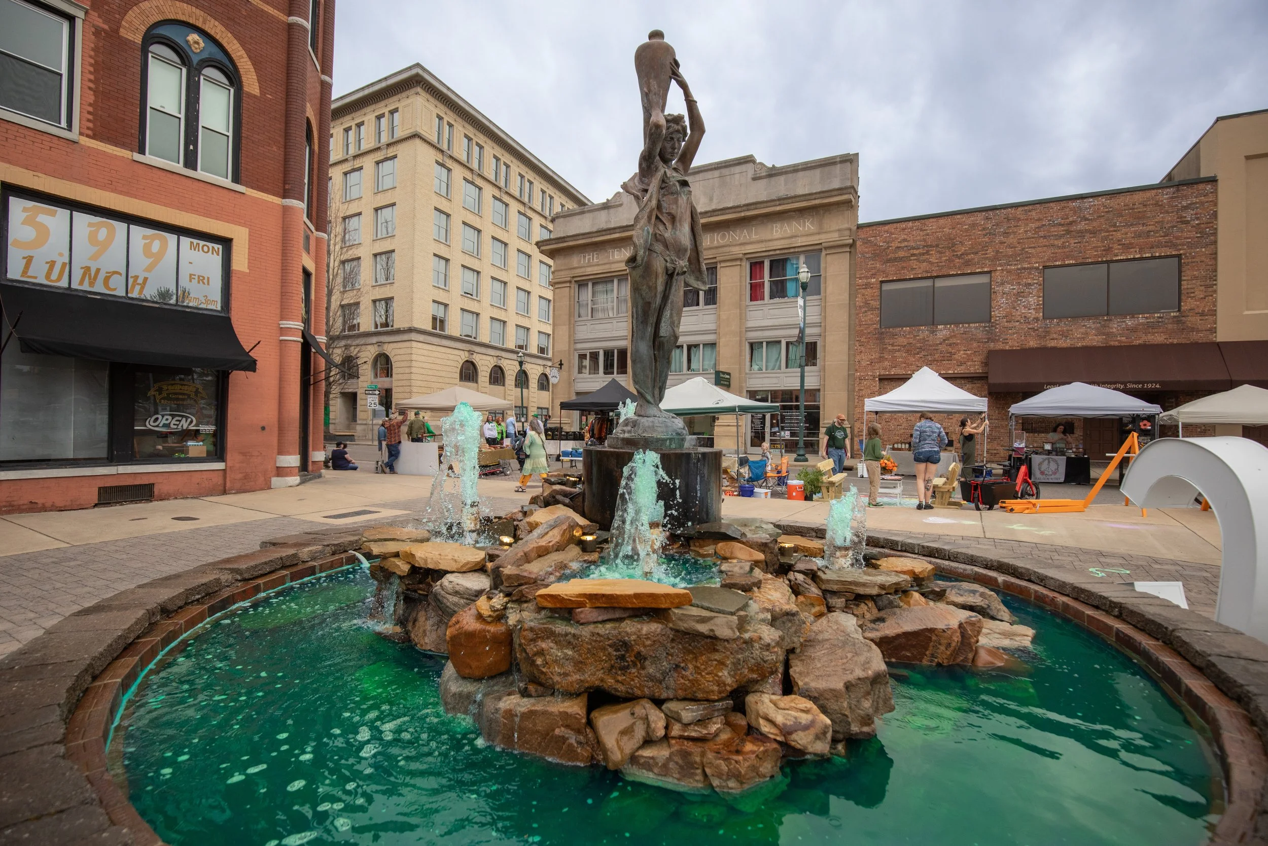 A statue of a woman holding a jar above her head stands in a fountain with water cascading over rocks, surrounded by an urban plaza with vendor booths and pedestrians.