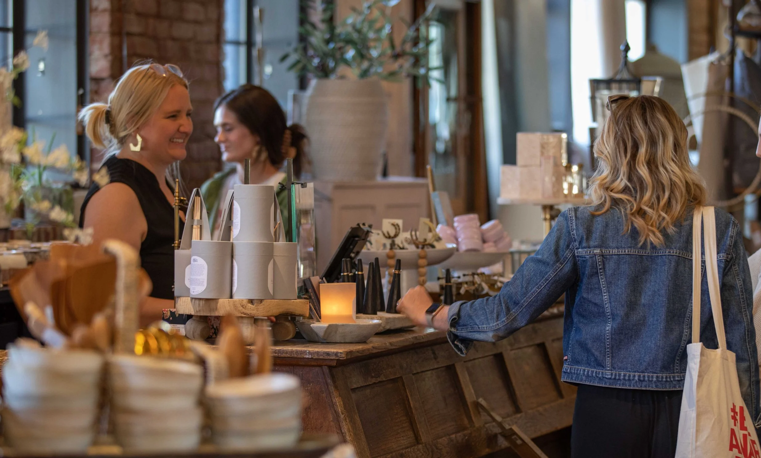 A retail shop with a woman customer at the counter talking to a smiling cashier. The shop has various decorative items and jewelry displayed on the counter and shelves.