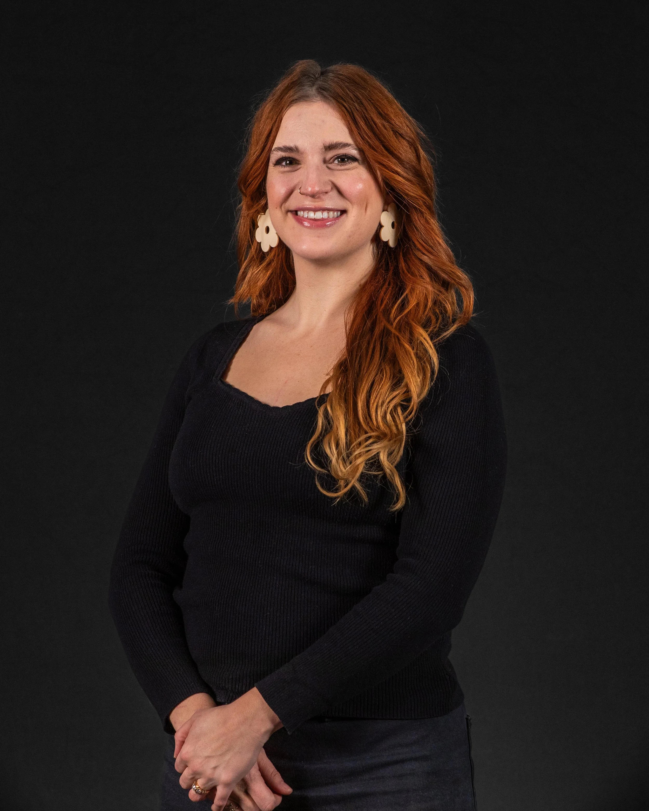 Portrait of a smiling woman with long red hair, wearing a black long-sleeve top and large white earrings, standing against a dark background.