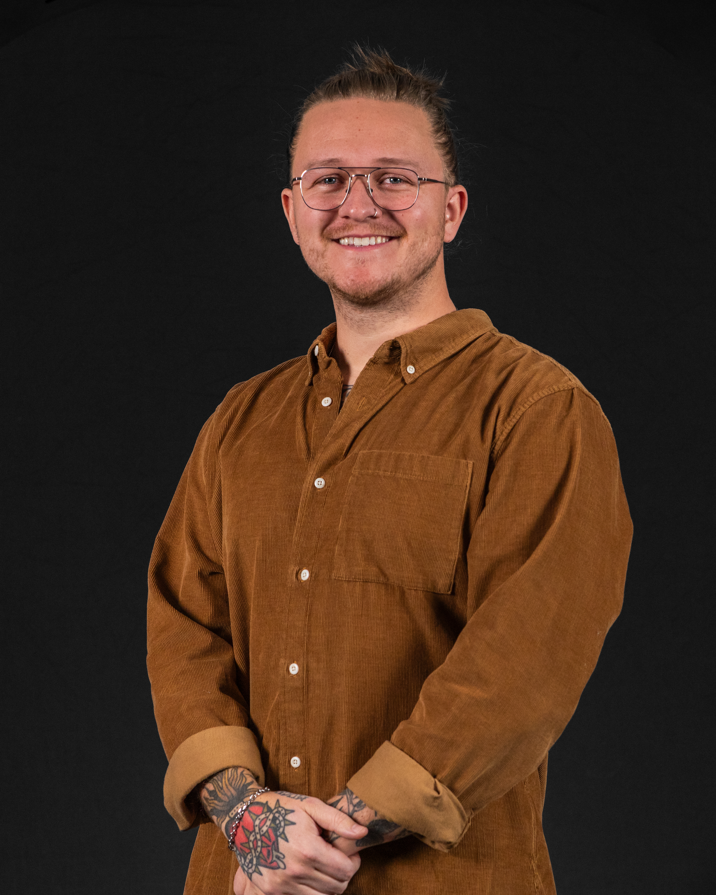 A smiling man with glasses and tattoos, wearing a brown button-up shirt, standing against a black background.