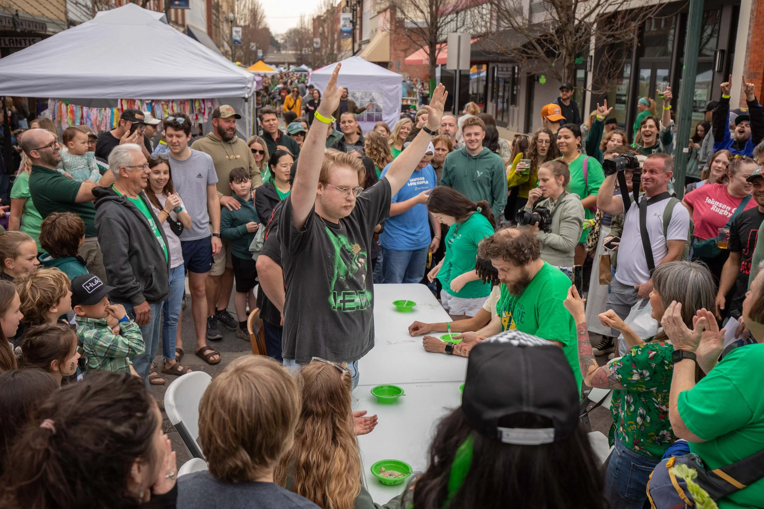 A large crowd gathered on a street during a festival, with some people raising their hands and taking photos. A person with glasses and red hair is seated at a table with green bowls, while others around are watching and cheering. There are tents and booths in the background, and people of various ages are participating.