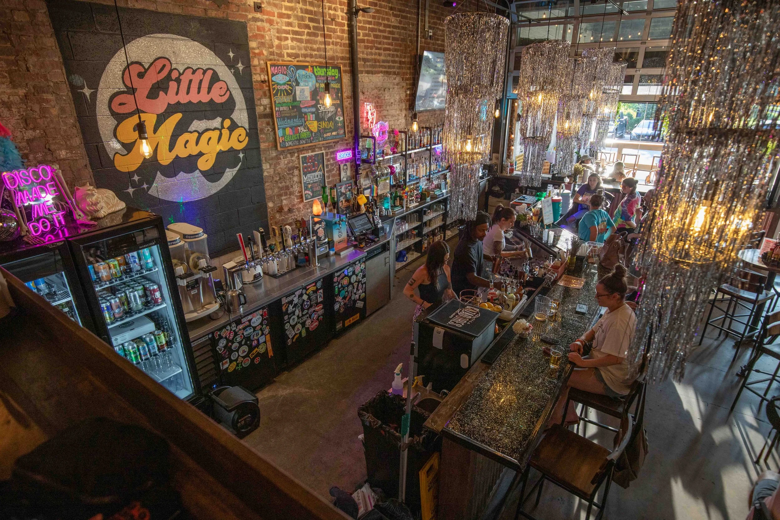 Interior of a lively bar with a brick wall featuring a "Little Magic" sign, bright neon lights, and shiny chandeliers. Several patrons are seated at the bar and tables.