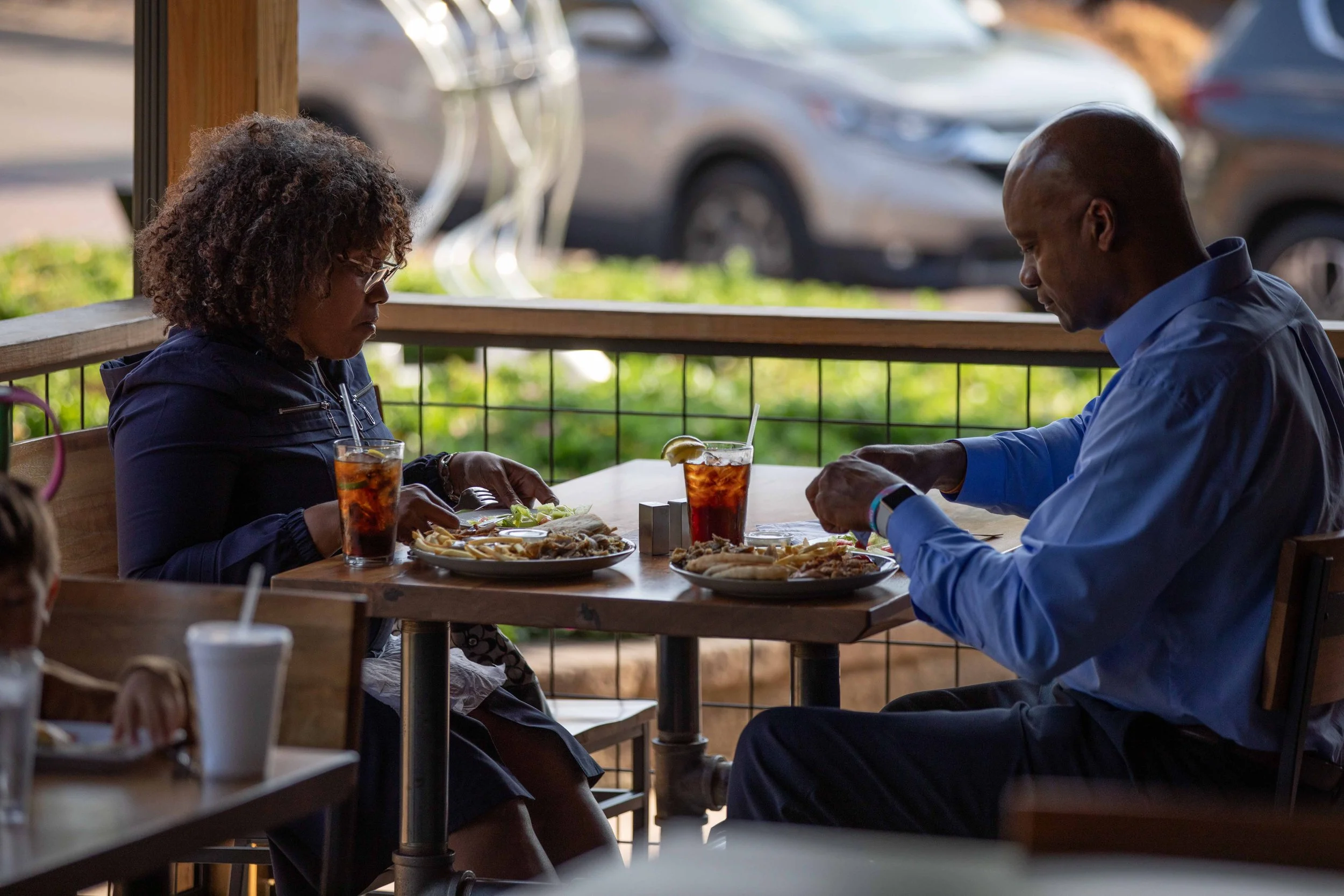 A man and a woman sitting at a wooden table outside a restaurant, looking down at their plates with food. The woman has curly hair and is wearing glasses, while the man has a shaved head. Two glasses of iced tea are on the table.