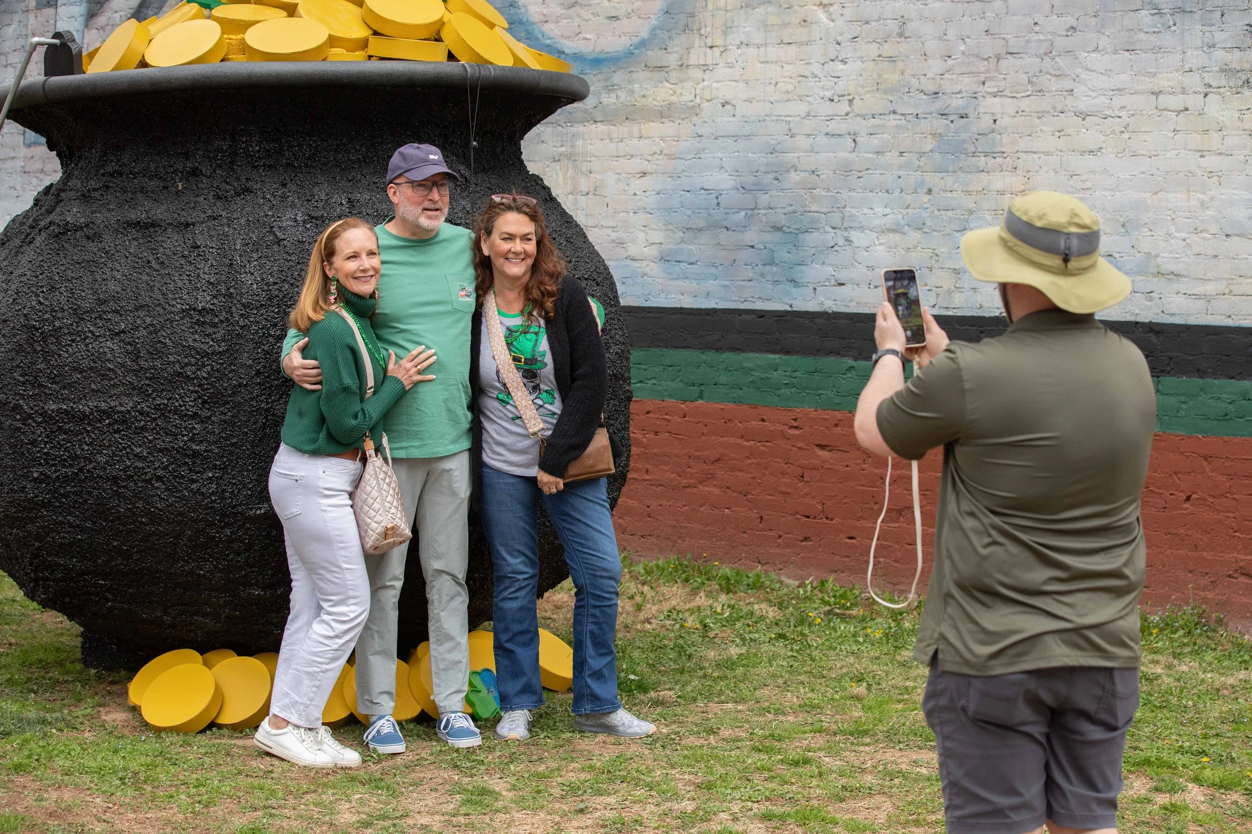 Three people are taking a photo in front of a large, colorful art installation of a pot filled with yellow coins. The group is standing on grass with the person on the right taking a photo using a smartphone.