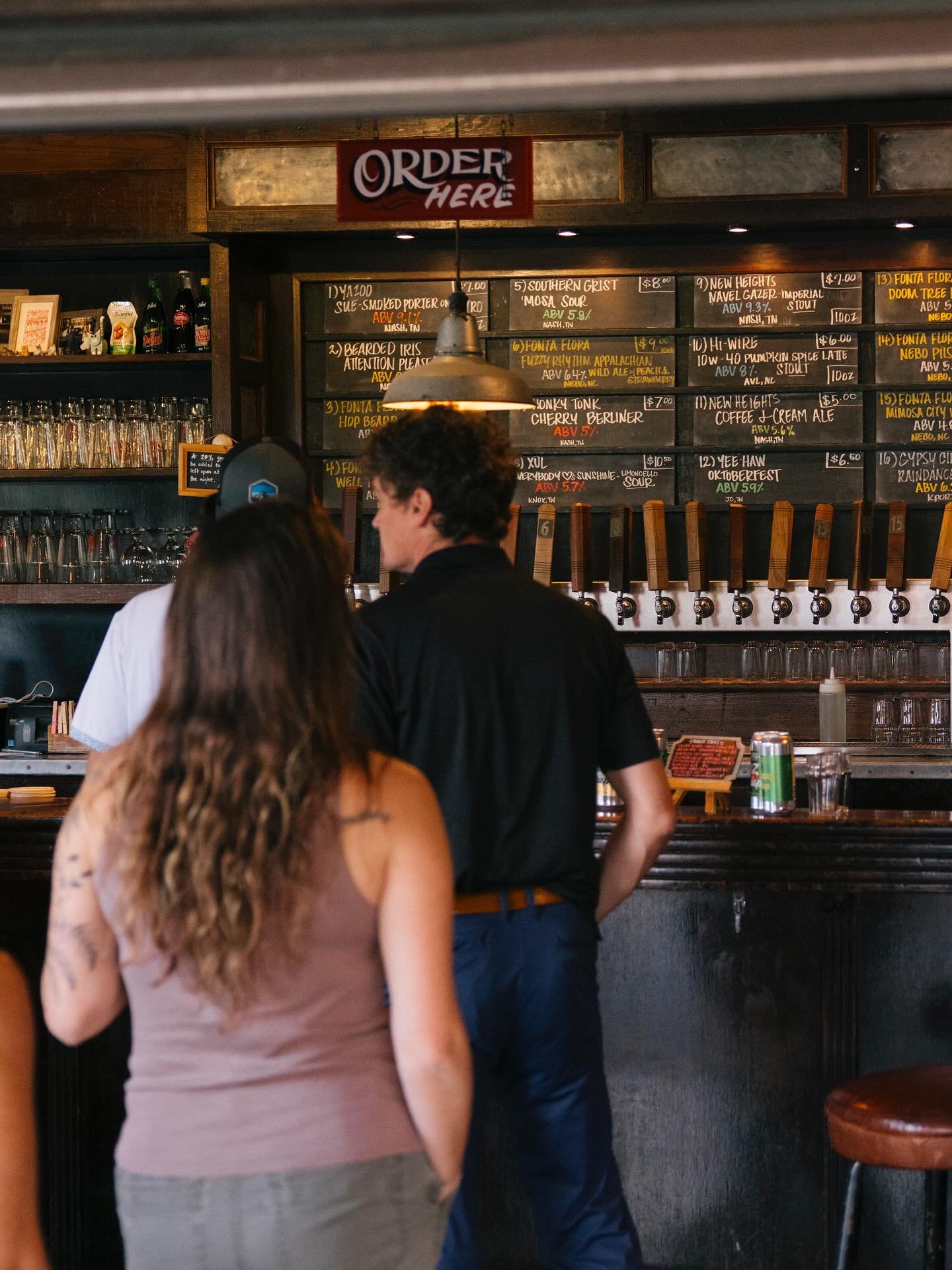 People ordering drinks at a bar, with a chalkboard menu behind them displaying various beverage options and prices.