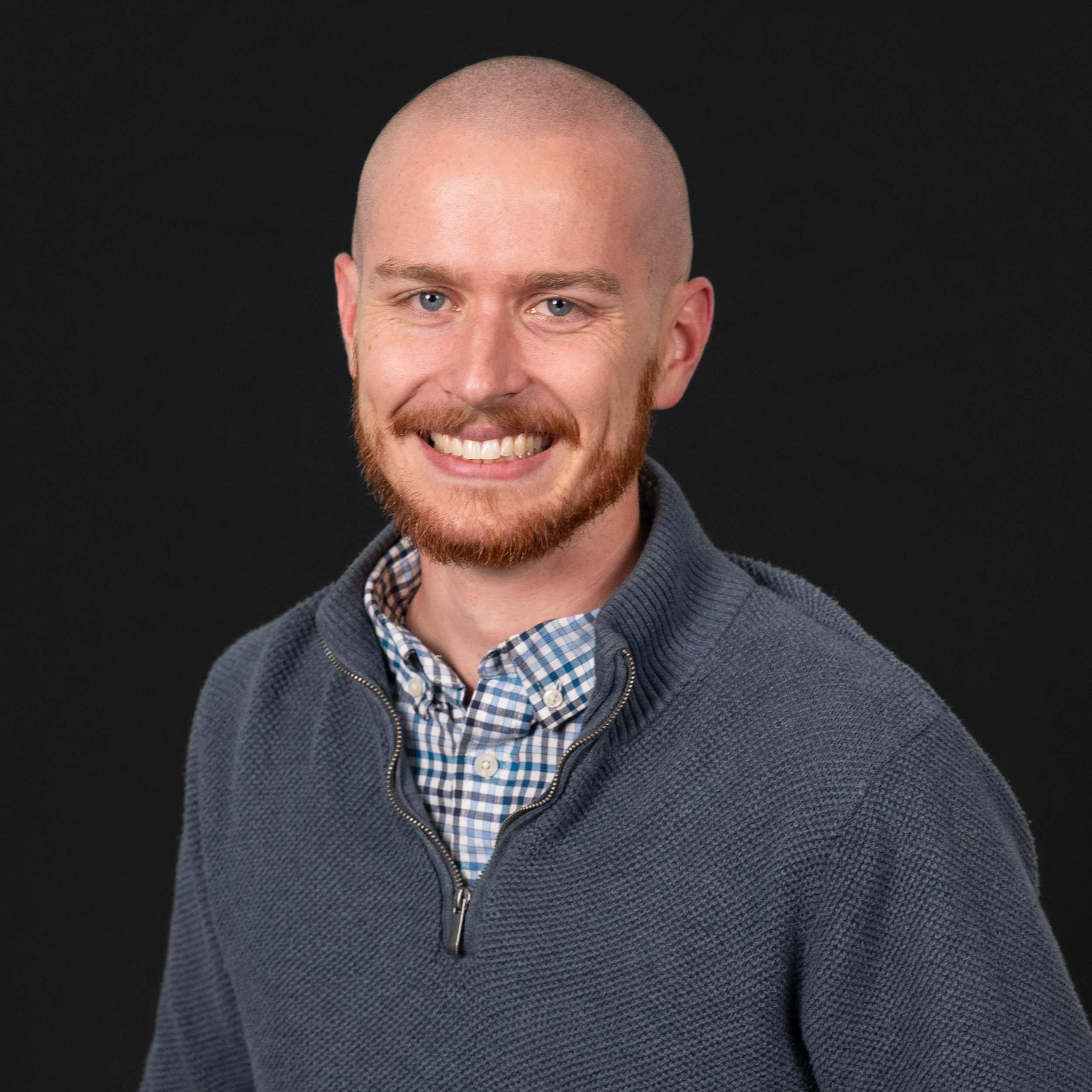 A smiling man with a beard and short hair, wearing a grey sweater over a checkered shirt, against a black background.