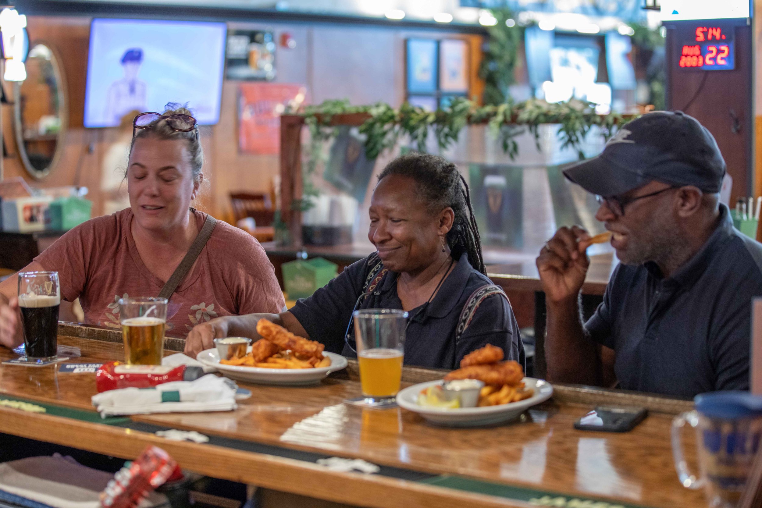 Three people sitting at a bar with plates of fried foods and glasses of beer, enjoying a meal together in a lively restaurant.