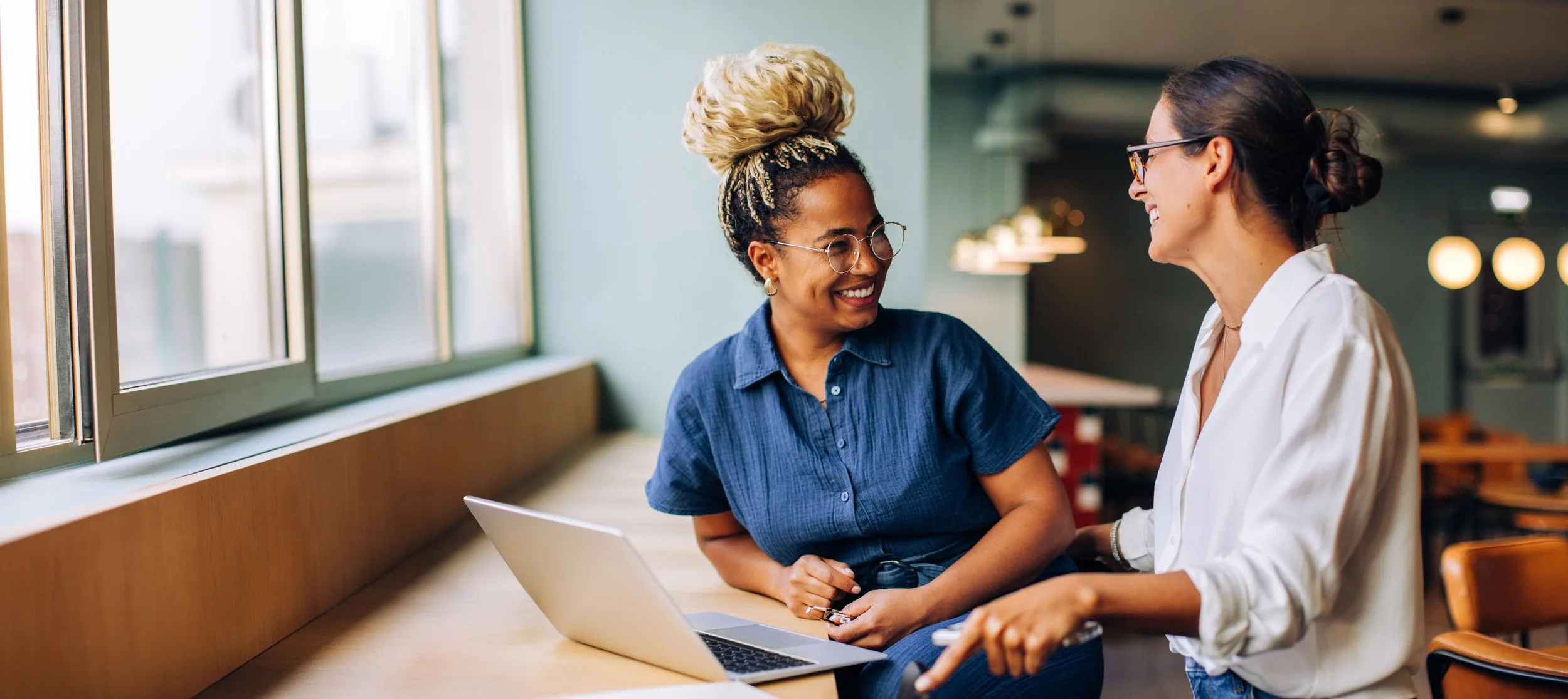 Two women smiling and talking in a cafe or office with a laptop on the table.