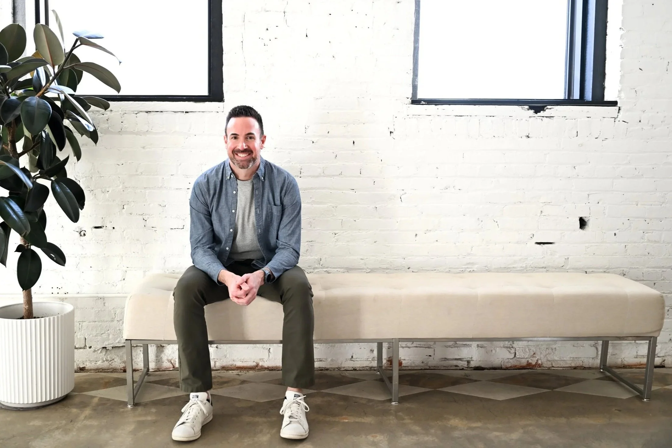 A man with a beard, wearing a blue shirt, gray pants, and white sneakers, sitting on a beige couch in a room with white brick walls and large windows, with a potted plant beside him.