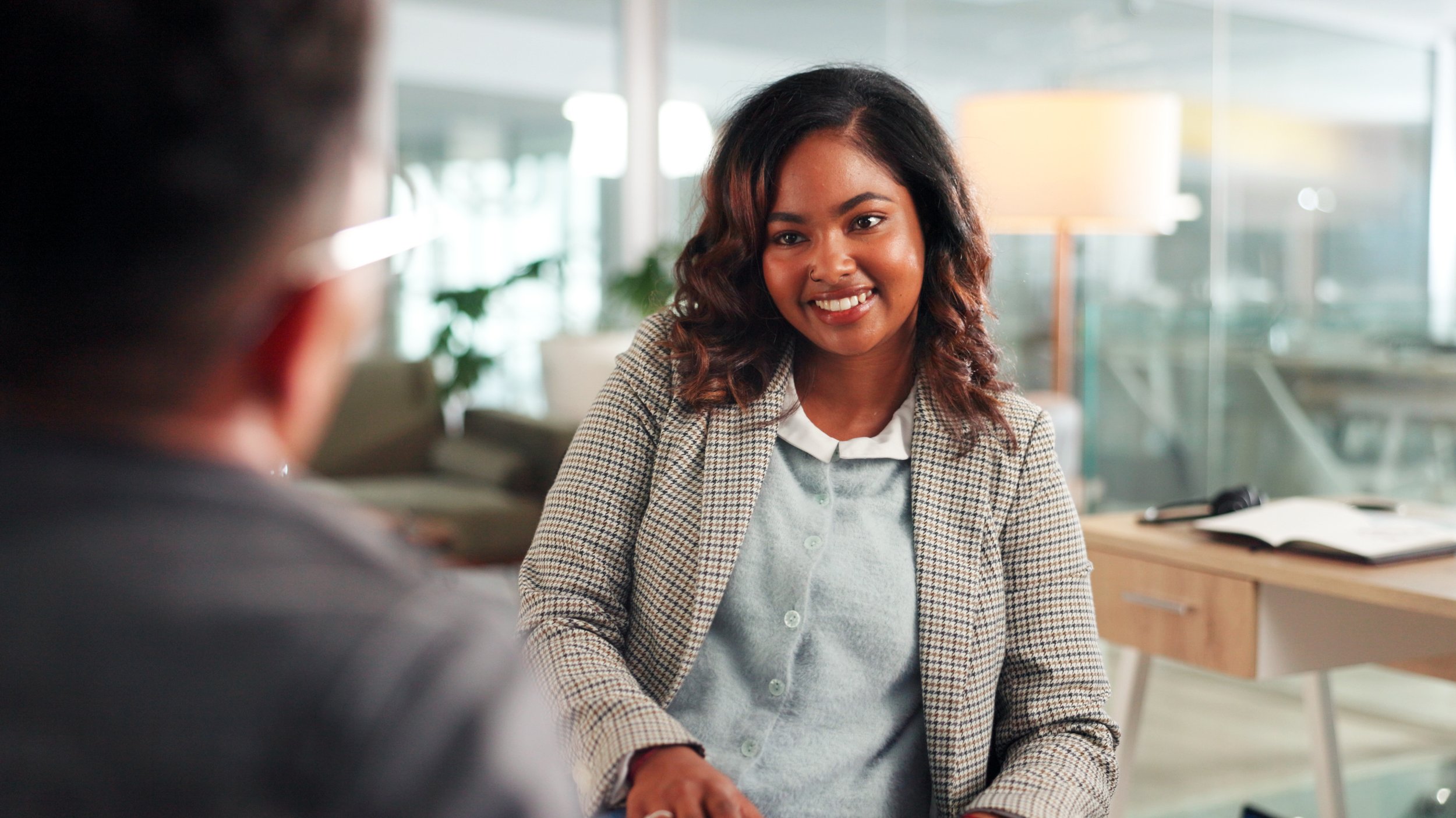 A woman with curly hair, wearing a checkered blazer and a light-colored shirt, smiling during a conversation in an office setting with a person whose back is visible in the foreground.