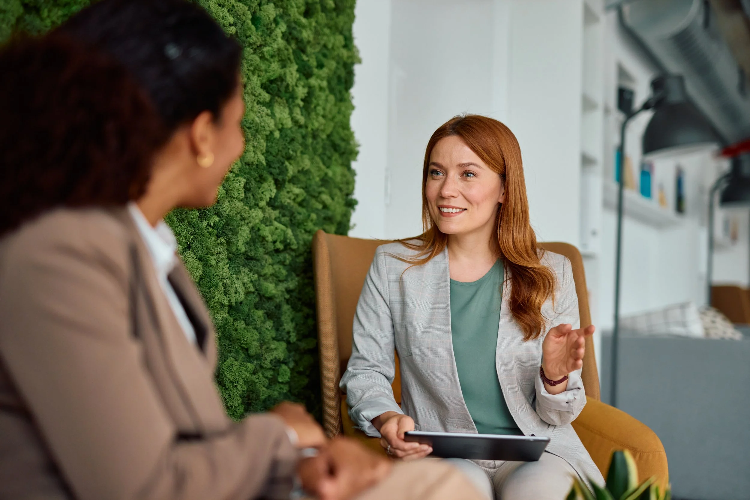 Two women engaged in conversation, one with dark hair facing away and the other with red hair smiling, sitting in a modern, bright office space with a green moss wall in the background.