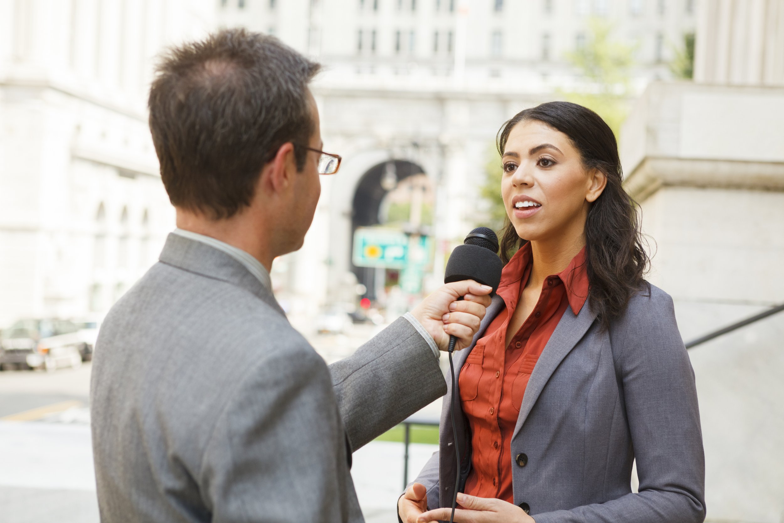 A woman being interviewed outdoors with a microphone held in front of her, man in gray suit facing her, cityscape background.
